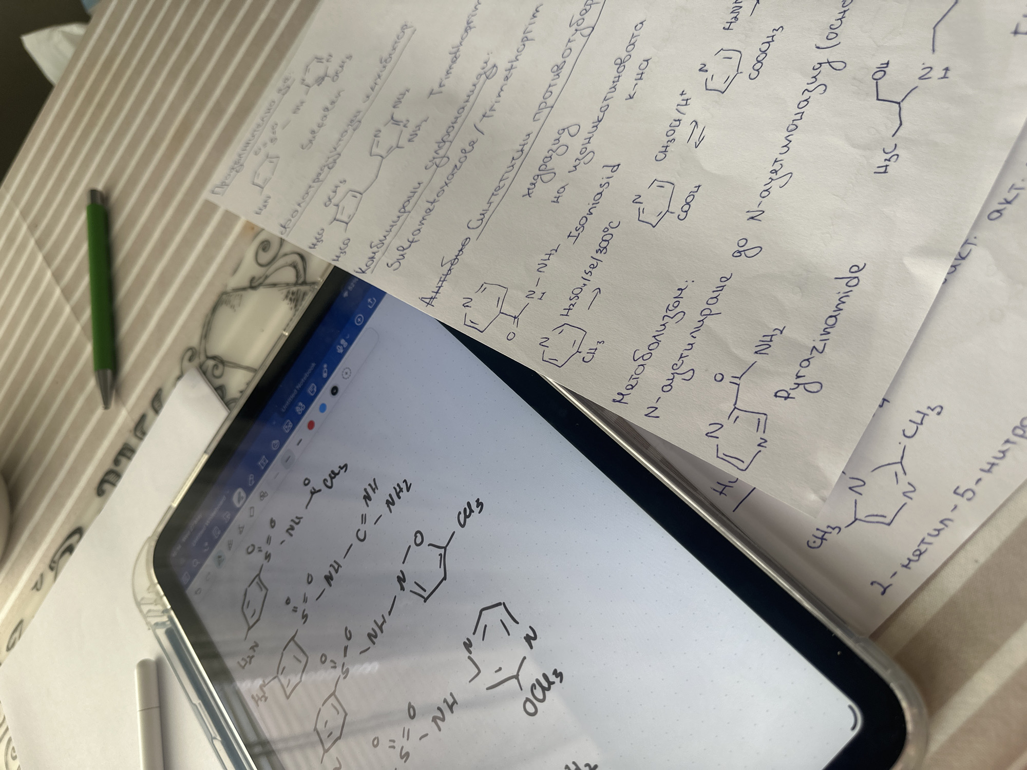 A close-up view captures a collection of handwritten notes and chemical structures scattered on a surface next to a tablet. The tablet displays drawn chemical structures on its screen. The notes contain chemical structures, chemical equations, and corresponding text, primarily focusing on chemistry-related topics. A green pen is also visible. The location is not discernible from the image.