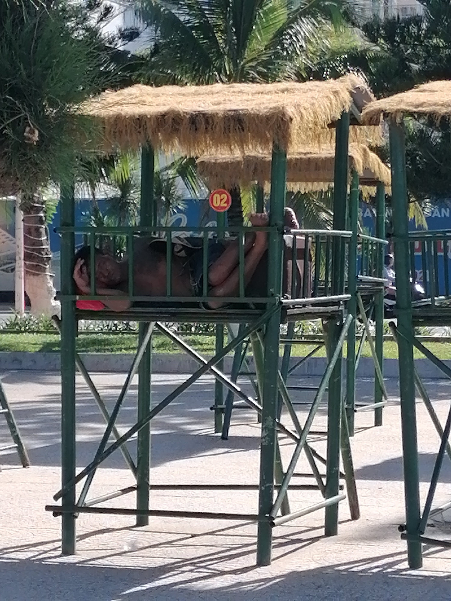 A man is sleeping on a raised, green metal structure with a thatched roof. The structure appears to be a lifeguard stand, given the presence of a red circular sign with the number "02" on it. It's one of several similar structures lined up in a row. Palm trees are visible in the background, along with parts of buildings. The setting is outdoors, and the ground is paved. The image was taken in the city of Nha Trang Ward, Vietnam.