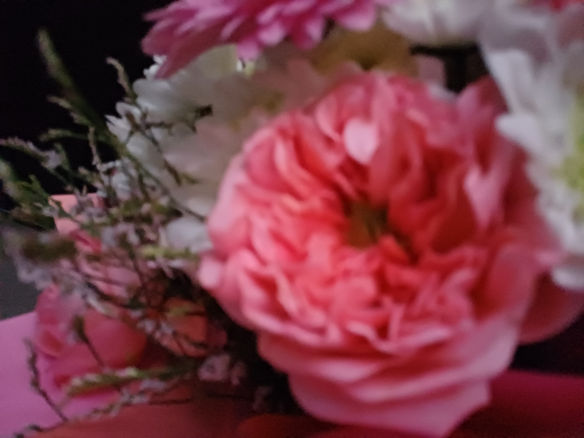 This is a close-up, blurred image of a floral arrangement. The primary focus is on a large, pink rose with many ruffled petals in the foreground. Behind it, other flowers are visible, including white and pink blossoms, along with greenery and delicate, wispy stems. The background is dark and indistinct. The image does not provide any information about a specific city or location.