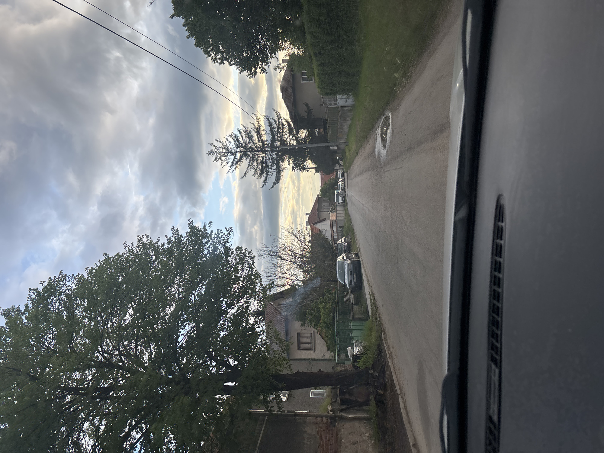 The view from inside a vehicle looking down a narrow road in Golema Rakovitsa, Bulgaria. Houses line the street, with cars parked along the side. Trees are visible on either side of the road. The sky is overcast with clouds.