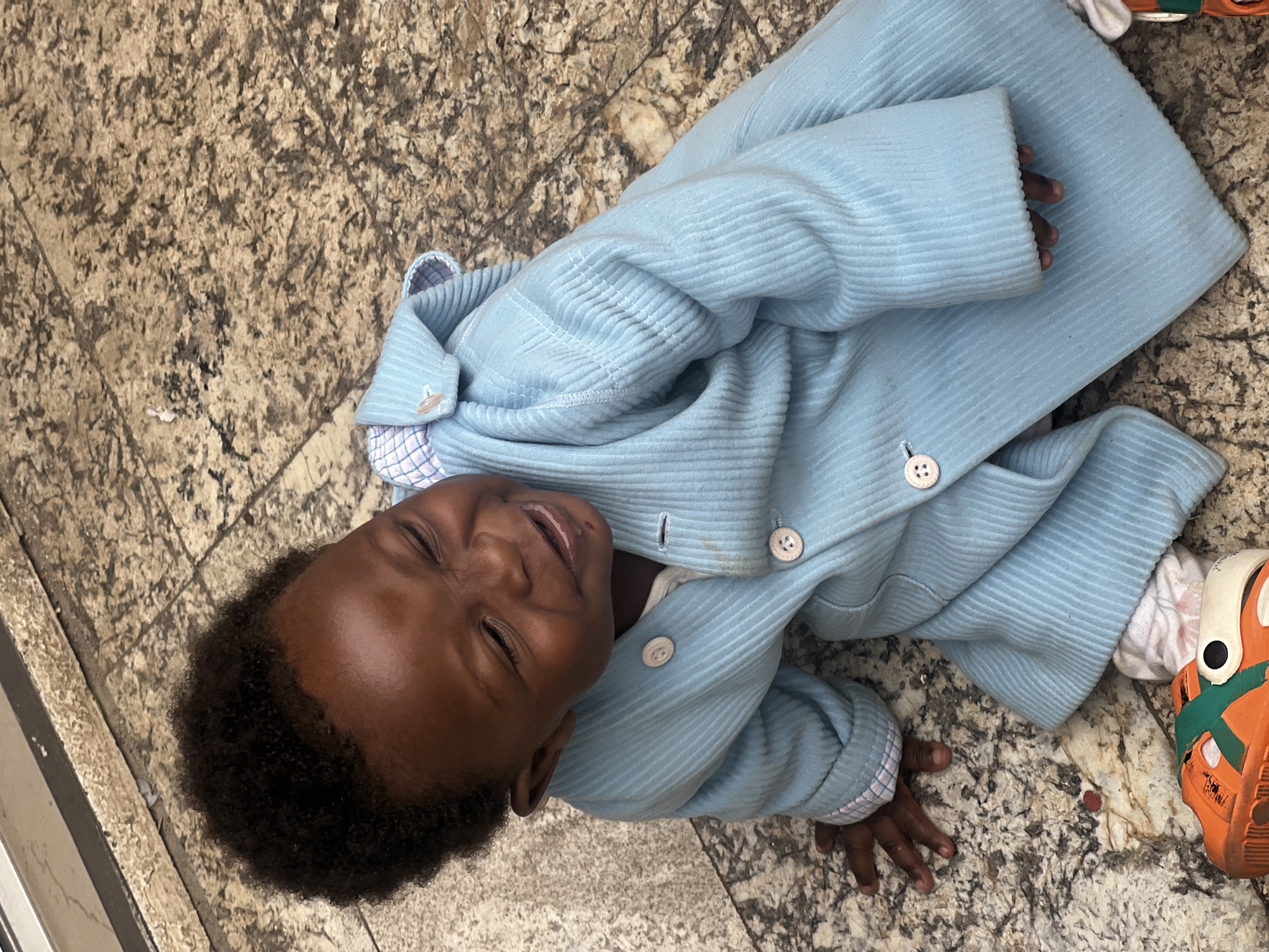 An overhead shot captures a child lying on a granite floor. The child, with dark hair and skin, smiles slightly. He is dressed in a light blue corduroy jacket with white buttons, and a glimpse of a checkered collar is visible. The child wears an orange and green shoe on one foot. The location is Gura Top, Nigeria.