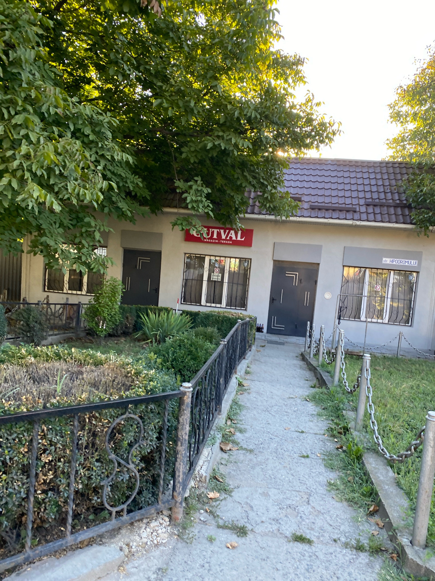 The image depicts a street-level view of a single-story building in Chișinău, Moldova. The building has a dark brown tiled roof and a light gray facade. A red sign with the text "UTVAL" is visible above a window. The building features at least three windows with bars and two dark gray doors. One window on the right has a sign that reads "str. HIPODROMULUI". The foreground shows a pathway leading to the building, bordered by a black wrought-iron fence on the left and a chain-link fence supported by metal posts on the right. Lush green trees with dense foliage overhang the building from the left. The area appears to be a commercial or service establishment, possibly a shop or cafe, with a small patch of green grass and landscaping. The lighting suggests it is daytime, with sunlight filtering through the trees.