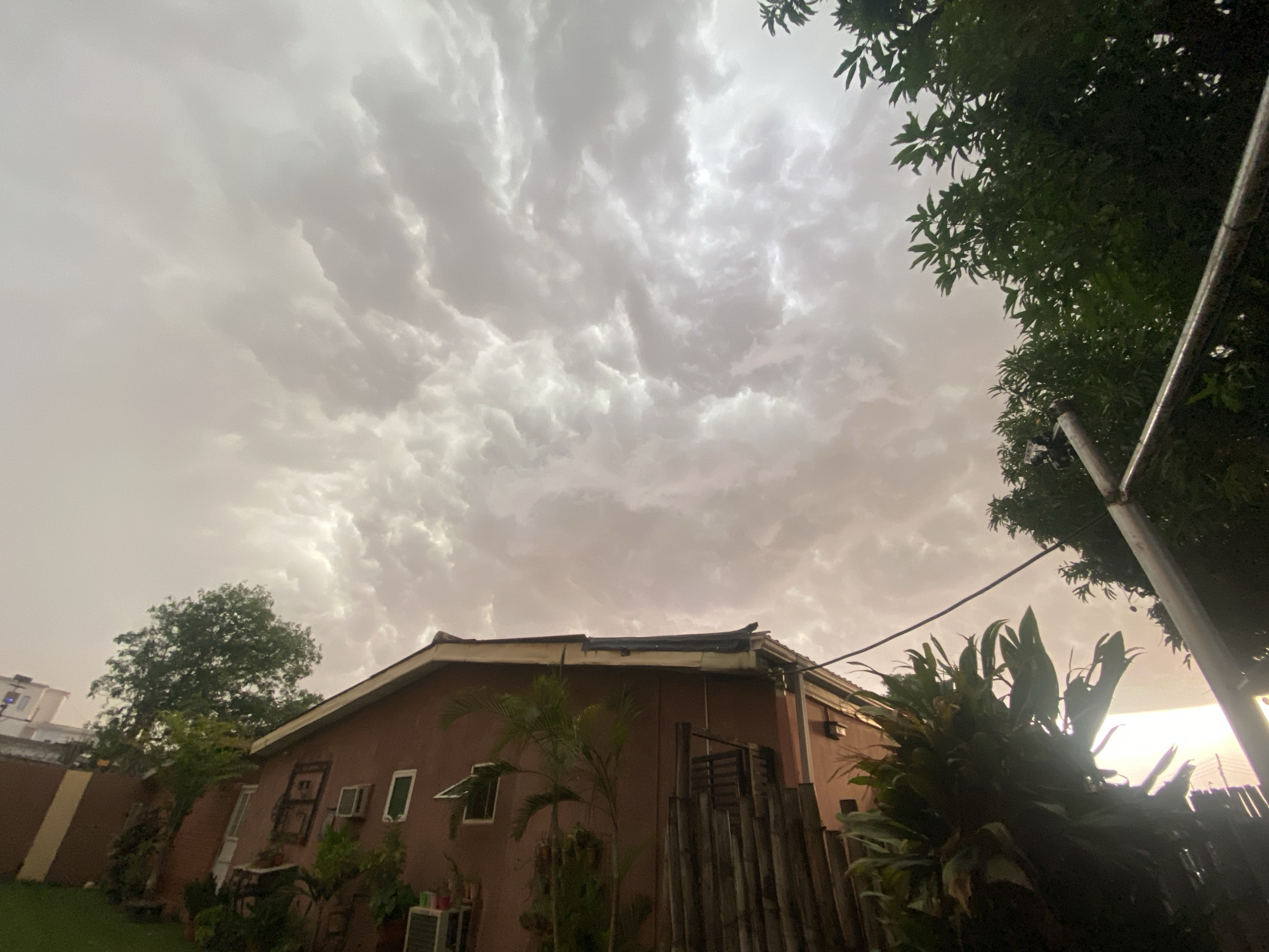 The image shows a view in Barden Yana, Nigeria. Overcast skies dominate the scene. A reddish-brown, single-story building is centered. Trees and dense foliage surround the building. A wooden fence partially obscures the building's facade. In the distance, a lighter-colored structure is visible. The overall lighting suggests it is either early morning or late evening. A pole is on the far right of the image. There are no people visible in the image.
