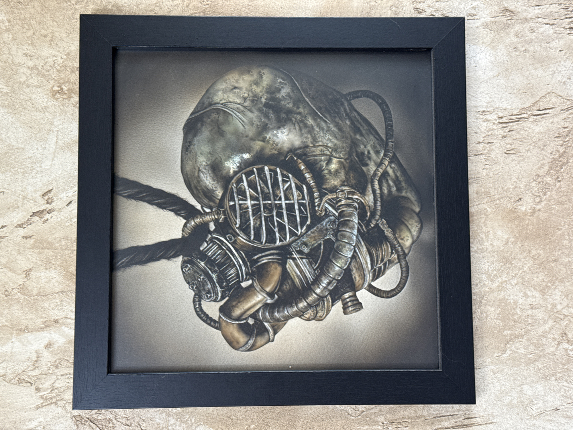 A framed print hangs against a textured wall. The black frame surrounds an image of a detailed, possibly metallic, mask or helmet resembling a distorted human head. The mask features intricate pipes, tubes, and a grated filter over the face area. The background of the print is a gradient of gray. The wall has a mottled, stone-like texture with varying shades of beige and brown.