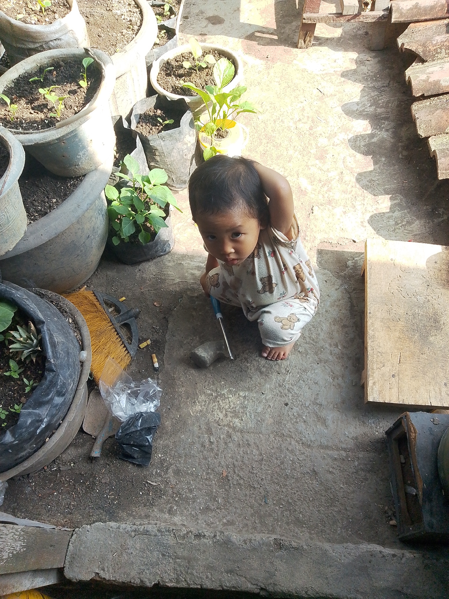 The image shows a young child, possibly a toddler, squatting on a concrete ground surrounded by potted plants and gardening tools. The child is wearing a light-colored outfit with a teddy bear pattern. To the left of the child is a large black plastic pot containing a plant, and next to it, a yellow-bristled broom and a small shovel. There are also several cigarette butts scattered on the ground. In the background, more potted plants are visible, some in large grey planters and others in black plastic bags. A wooden plank is placed to the right of the child. The setting appears to be an outdoor space, likely a yard or a balcony, in what could be a residential area in Bandar Lampung, Indonesia, given the typical urban landscape and vegetation observed. The overall scene suggests a moment of play or exploration by the child amidst gardening activities.