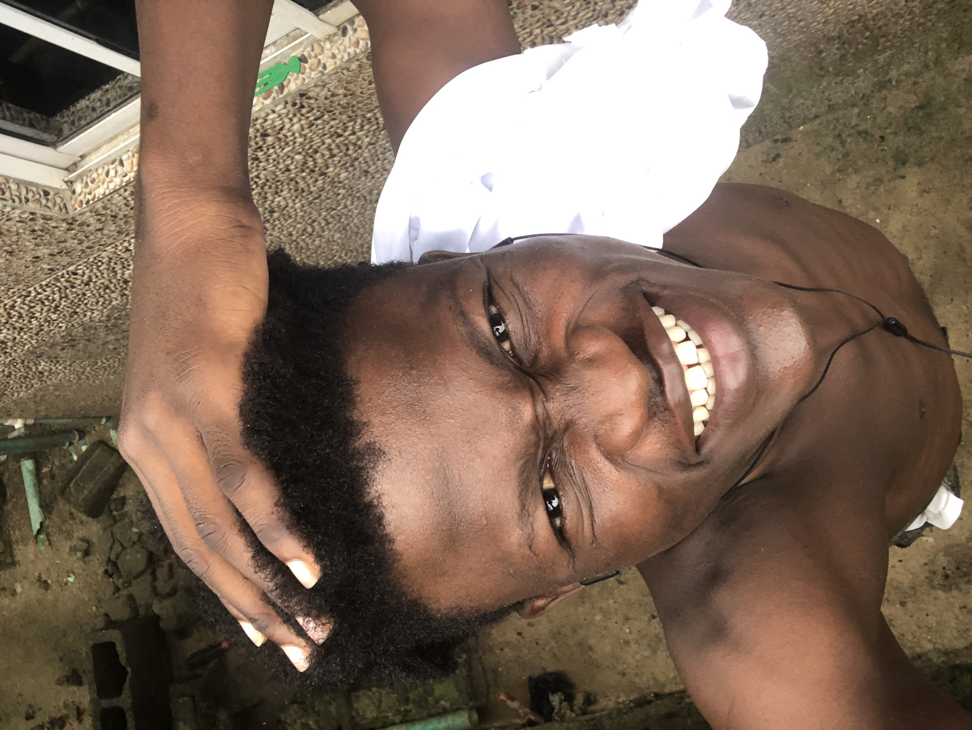 A high-angle, medium close-up shot captures a young Black male from the chest up, his head tilted back and eyes scrunched shut as he grins widely, revealing his teeth. His right hand is on top of his head, fingers splayed, touching his short, dark, curly hair. He is wearing black earbuds, with wires visible around his neck and chest. A piece of white fabric is draped over his left shoulder. The background suggests an outdoor, possibly urban, environment. To the left, the ground appears to be concrete or dirt, with scattered debris including what looks like a broken cinder block and some piping. To the right, a textured wall composed of small, multi-colored pebbles runs alongside a white-framed window with vertical panes. The lighting is natural and appears to be daylight.