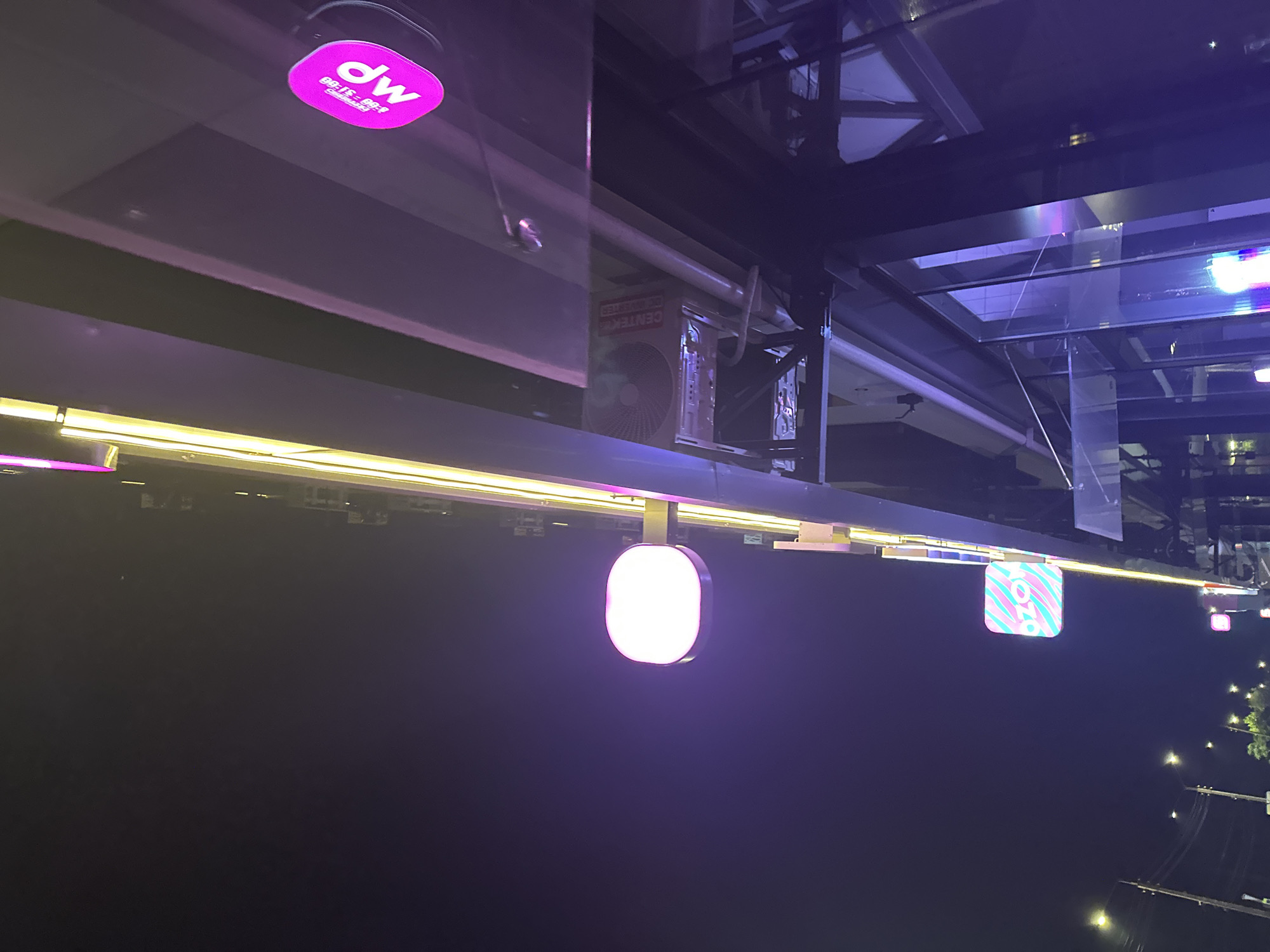 The image is a night-time photograph taken from a low angle, looking up at a building facade illuminated by neon lights. The building appears to be a commercial establishment, possibly a store or restaurant, located in the city of Nevinnomyssk, Russia.
Prominently displayed are several illuminated signs. On the right side of the image, a bright pink oval sign with white lettering is visible. The lettering is partially obscured but appears to read "MP" with some smaller text above it. Further to the left, a square sign with a diagonal striped pattern in pink and turquoise colors is lit up. The word "ozon" is discernible in white text on this sign. Another rectangular sign with a pink outline and white illumination is visible lower down and to the left.
The building itself features a modern architectural design with large glass windows and dark framing. Above the glass facade, a series of horizontal beams are illuminated by yellow-toned lights, creating a vibrant effect against the dark night sky. An air conditioning unit is mounted on the exterior wall. Streetlights are visible in the background, indicating an urban setting. The overall ambiance is one of a lively, illuminated commercial district at night.