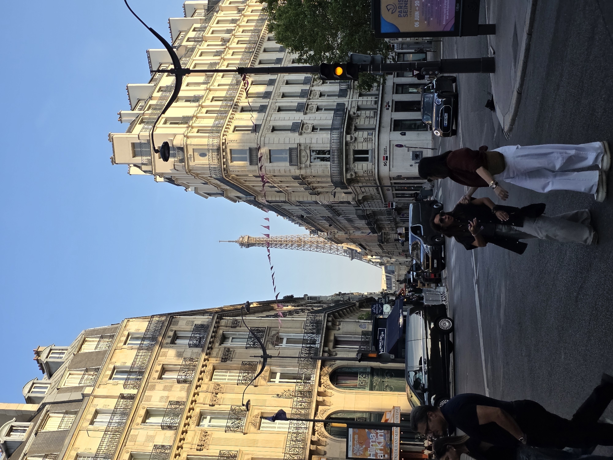This media file captures a street scene in Paris, France, with a clear view of the Eiffel Tower in the distance. The perspective is from street level, looking up at ornate Haussmannian buildings lining a boulevard. People are visible walking on the sidewalk and street. One group appears to be posing for a photo or interacting, with one individual standing and two others in the foreground leaning down. A few cars are parked or driving on the street, including a black Mini Cooper.
The time of day appears to be late afternoon or early evening, indicated by the warm, golden light illuminating the buildings and the clear, blue sky. The weather seems pleasant.
Visible text includes advertisements and signage. One sign mentions "PARIS SEINE L'ETE PARISIES" with dates "06 JUIN 20". Another sign advertises "Hilma af Klint" and "RESTAURANT SERVI". A street sign reads "MONCEAU". The Eiffel Tower stands prominently in the background, a recognizable landmark of Paris. The overall mood is a snapshot of daily life in a beautiful, iconic European city.