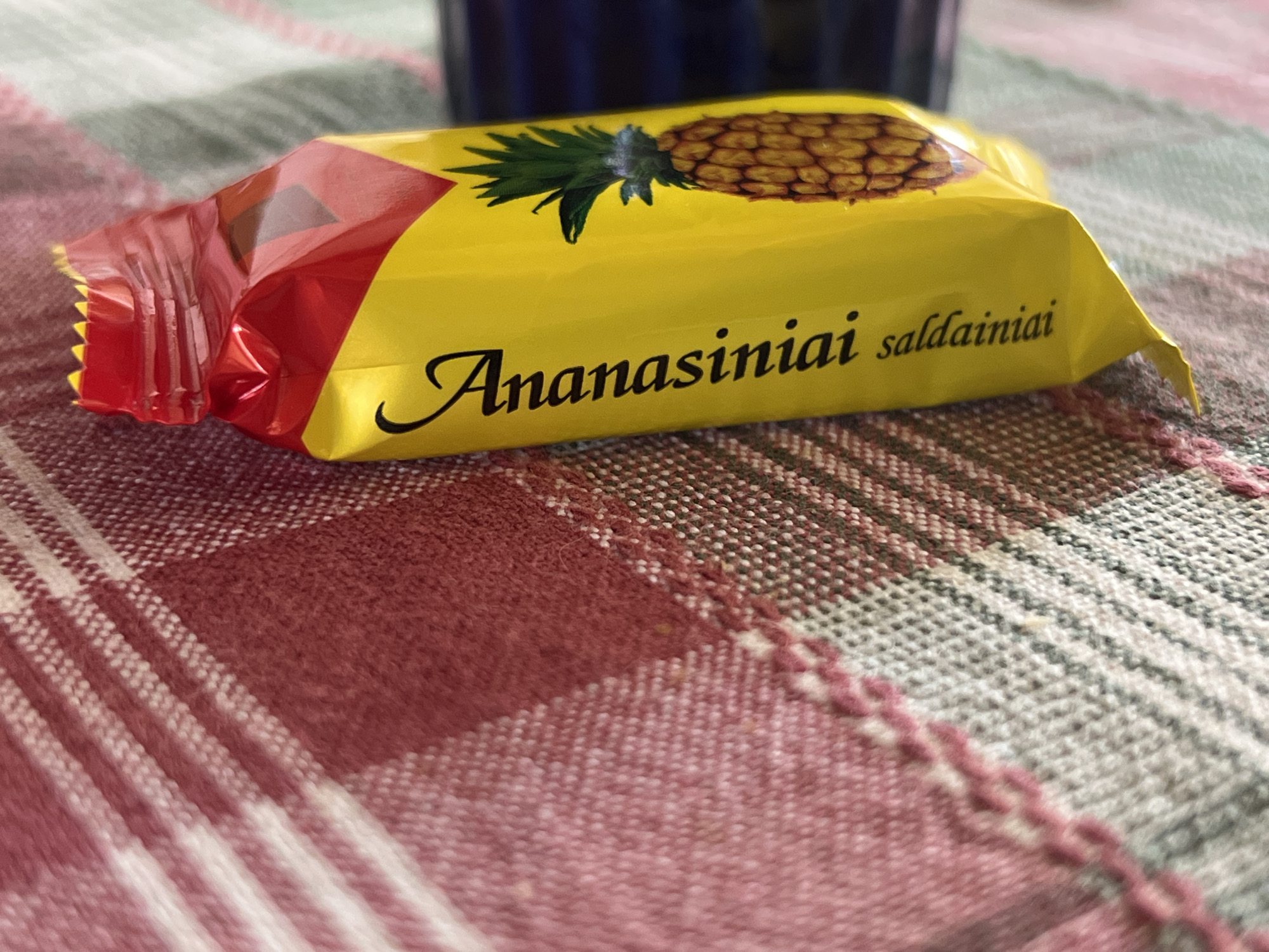The image shows a single, unwrapped candy with yellow and red packaging, featuring a picture of a pineapple and the text "Ananasiniai saldainiai". The candy is lying on a woven tablecloth with a plaid pattern of red, white, and gray stripes. The lighting suggests an indoor setting. The exact location in Daulėnai, Lithuania, is not discernible from the image alone.