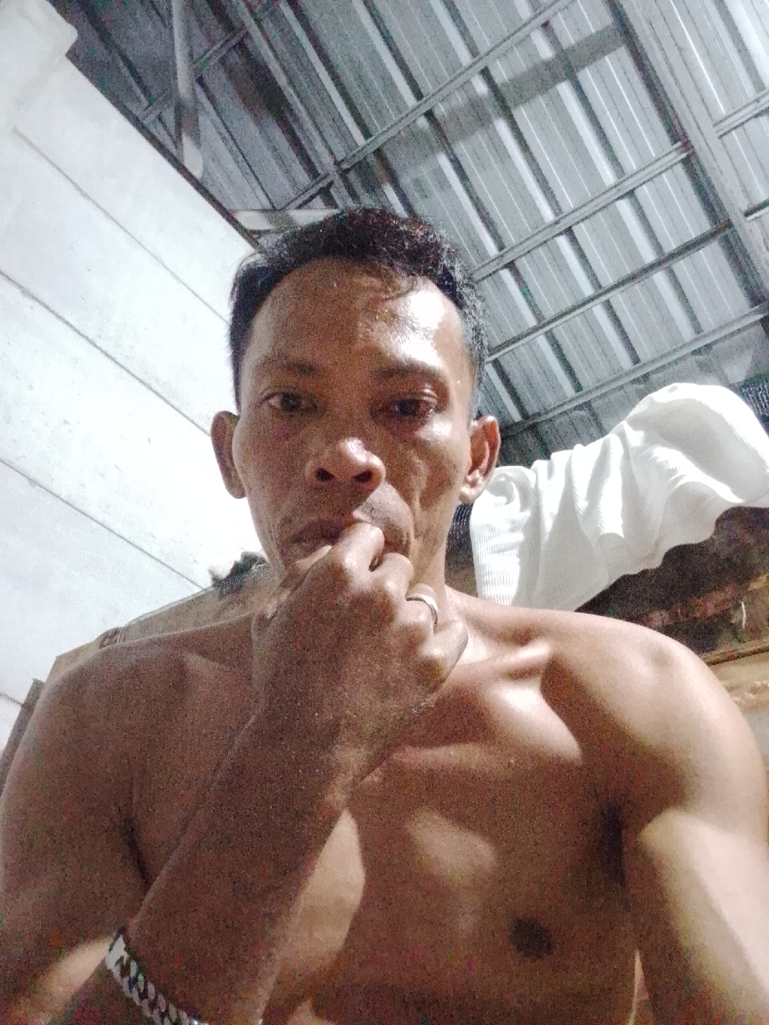 The photograph depicts a shirtless man indoors. He is looking at the camera with his fingers near his mouth. He is wearing a ring on his left hand and a bracelet on his left wrist. A white cloth is draped behind him. The location may be Bukit Ketok, Indonesia, but there is no clear corroborating evidence visible in the photograph to confirm this. The interior has a corrugated metal ceiling.