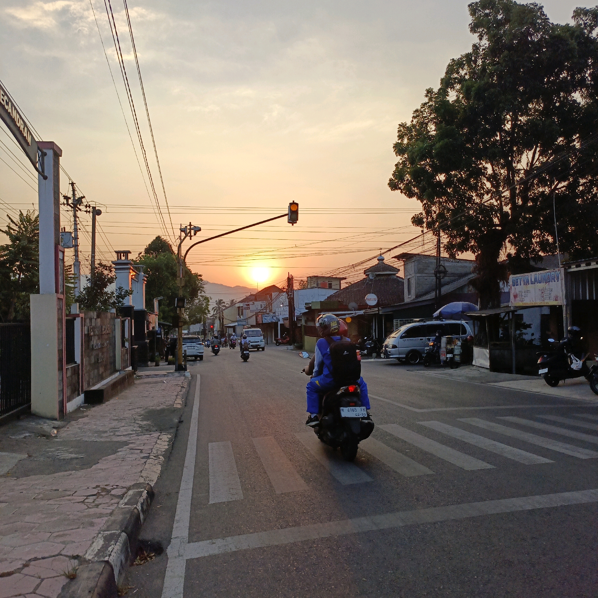 The image depicts a street scene in Salatiga, Indonesia, at sunset. The sky is a warm orange and yellow, with a bright sun low on the horizon, casting long shadows. A traffic light is illuminated amber. Several vehicles are present, including motorcycles and cars. Two people are riding on a motorcycle in the foreground, crossing a pedestrian zebra crossing. The street is lined with buildings, some appearing to be businesses, such as a laundry service. A large tree with dense foliage stands on the right side of the image. Utility poles and overhead wires are visible, typical of urban Indonesian infrastructure. The street has a paved surface with white markings for the pedestrian crossing.