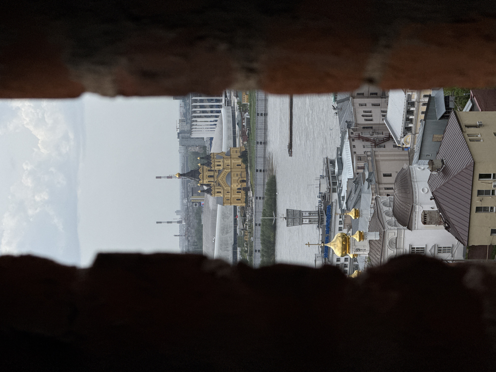 The image depicts a view of the cityscape of Nizhny Novgorod, Russia, as seen through a narrow opening in a brick structure, possibly a fortification. The buildings are a mix of architectural styles, including structures with golden domes and a large, yellow structure with green roofs. A wide river runs through the city, reflecting the sky. Industrial chimneys rise from the horizon, hinting at nearby industrial activity. The brickwork in the foreground is weathered and dark, contrasting with the brighter scene beyond. The image is slightly skewed, which is likely due to the angle of the viewpoint or image capture.