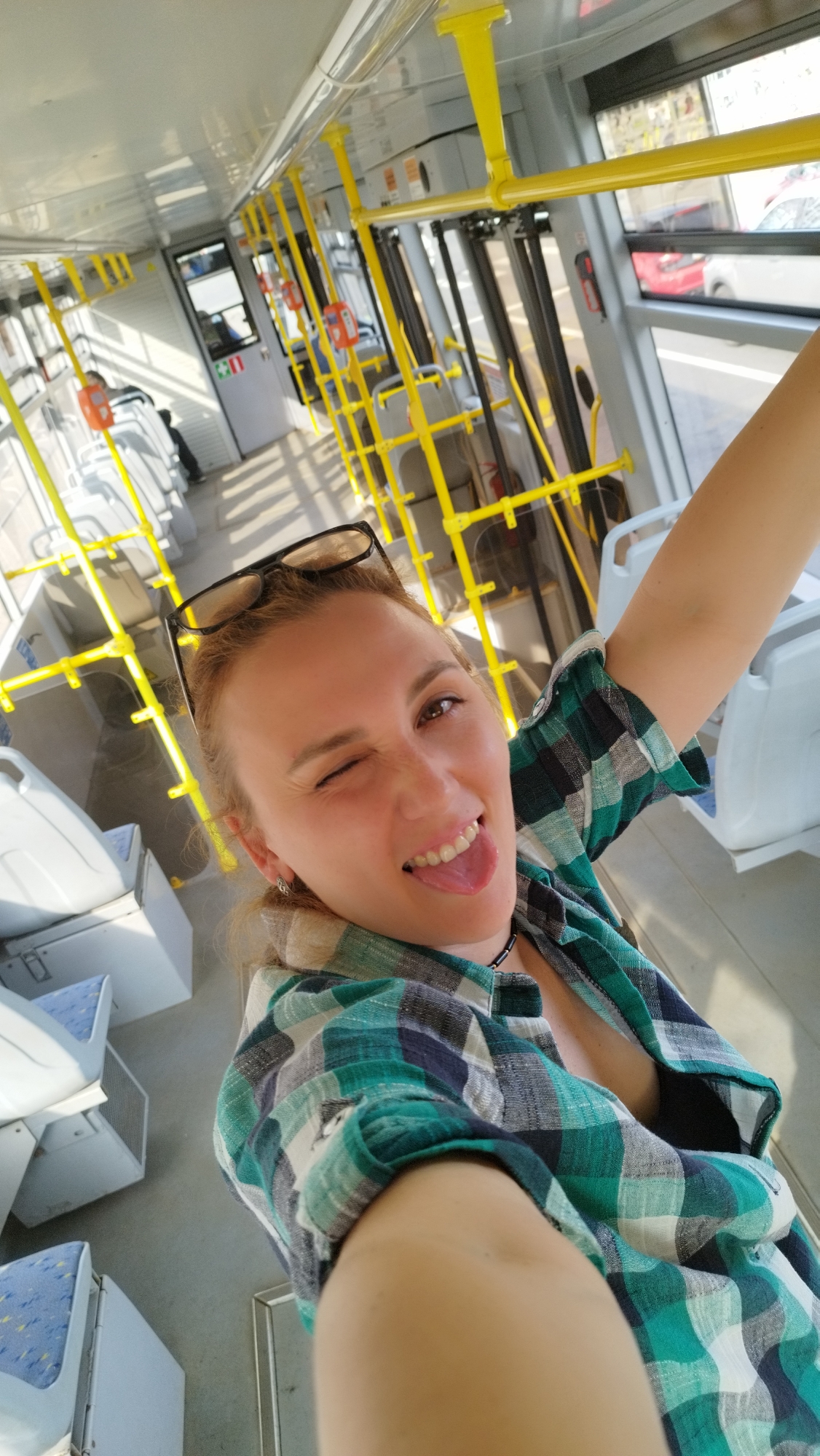 A woman with blonde hair tied back and wearing glasses on her head is taking a selfie inside a modern tram in Saint Petersburg, Russia. She is winking and sticking out her tongue, wearing a green and blue plaid shirt and a dark necklace. The tram interior features yellow handrails and poles, white seats with blue accents, and large windows showing a blurred urban scene outside, including another tram and buildings. The lighting suggests it is daytime.