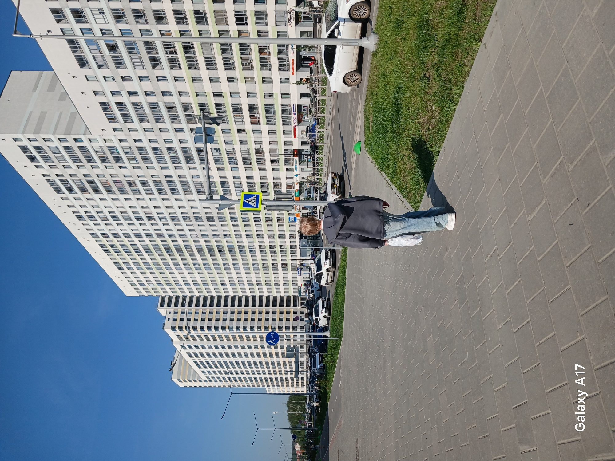 A vertical outdoor shot captures a person walking on a paved sidewalk, with a high-rise apartment complex in the background under a clear blue sky.
The person, seen from the rear, is wearing a dark grey jacket, blue jeans, and white sneakers. They are walking along the sidewalk, casting a shadow to their right.
The sidewalk is made of grey rectangular pavers. Adjacent to the sidewalk is a strip of green grass.
In the background, a large, white apartment complex dominates the view. The building is multi-story with numerous windows, creating a grid-like pattern. Streetlights are visible alongside the building. The photo appears to have been taken in the city of Yekaterinburg, Russia.
Vehicles are parked along the side of the road running between the apartment complex and the sidewalk. Several road signs are visible near the person.