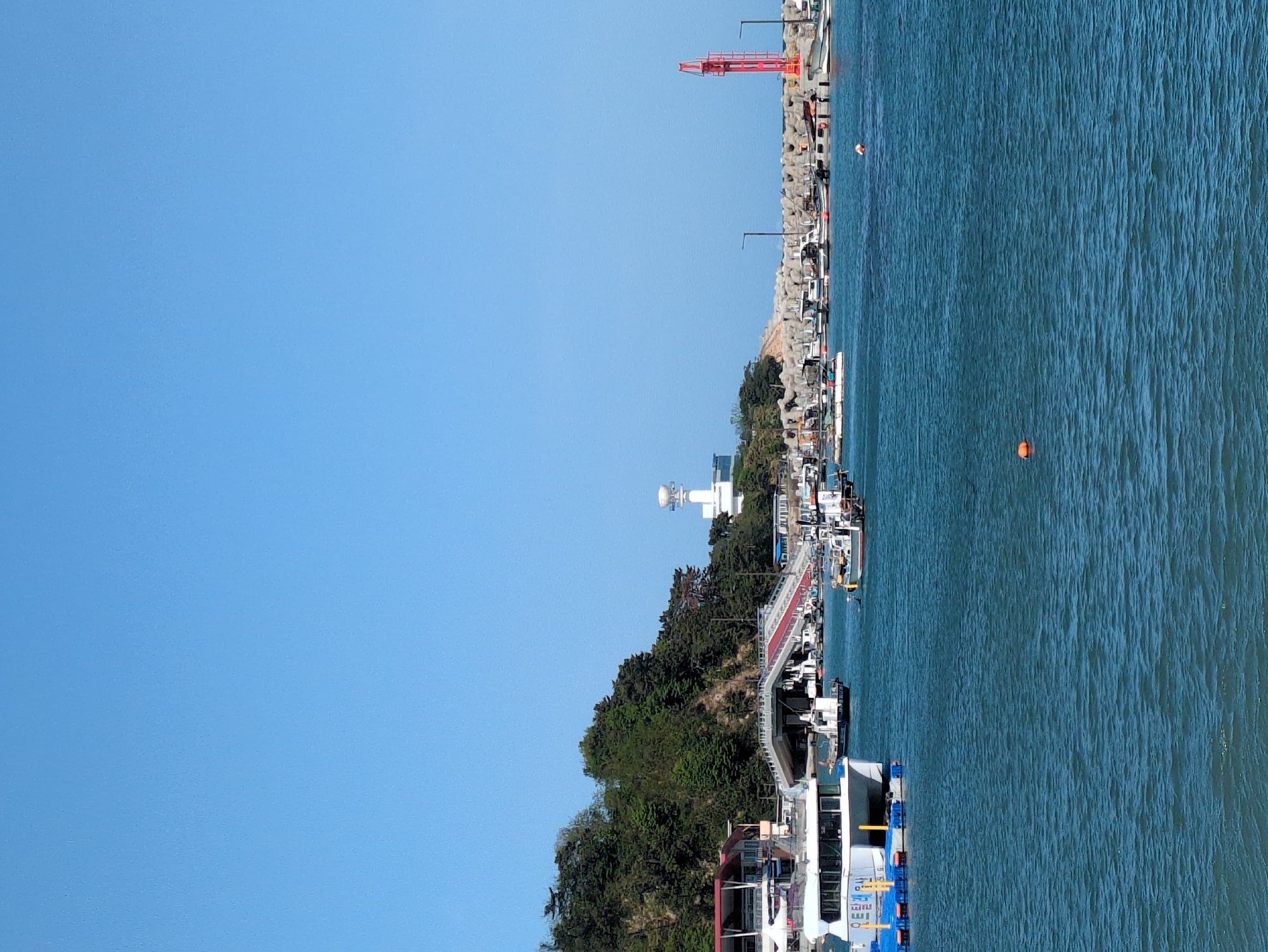 This outdoor scene depicts a harbor setting in Ulsan, South Korea, on a clear day under a bright blue sky. The focal point is the harbor's edge, where several boats are docked. A mix of greenery, including trees and shrubs, runs along the coastline, situated atop what appears to be a gradual slope or hill.
Notable features include a white structure with a cylindrical top, possibly a small tower or navigational aid, situated amongst the trees. There is a red pedestrian bridge stretching from the boat docks to the coastline. Large tetrapods protect the edge of the harbor. A red construction crane is visible in the distance at the harbor. There is at least one orange buoy in the water.