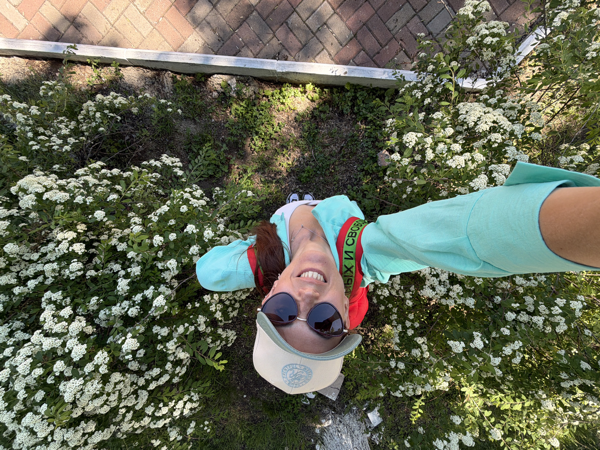A woman is captured from a high angle, looking upwards from what appears to be the ground. She is wearing a light blue top, a light-colored baseball cap, and sunglasses. A red and green band with text is visible on her upper arm. She is smiling broadly, with her teeth showing. The background consists of lush green bushes covered in small white flowers, with a paved area made of reddish-brown bricks visible on the right side. The presence of flowering bushes and paved walkway suggests an outdoor setting, possibly a park or garden area in Birobidzhan, Russia.