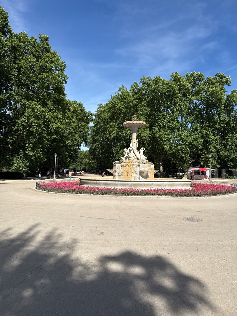The image displays a bright, sunny day in a large, verdant park, identified as being in Madrid, Spain. The central feature is an elaborate, multi-tiered stone fountain, possibly in a classical or allegorical style, with sculptural figures, including what appear to be nudes or cherubs at its base, and a smaller decorative element or bird at its very top. This fountain is encircled by a wide, shallow basin, which is further encompassed by a circular flower bed bursting with vibrant pink and red flowers.
The park is densely populated with mature, large-leafed green trees that frame the scene on both sides, their canopies reaching high into a clear, brilliant blue sky with only a few faint, wispy clouds. The sun is high, casting prominent, dark shadows of the tree foliage onto the light-colored paved ground in the foreground, suggesting it is mid-morning to early afternoon.
In the background, several indistinct figures of people are visible, moving or resting under the trees, adding a sense of casual activity to the park setting. To the far right, behind the fountain, a small red and white tent-like kiosk can be seen, possibly a refreshment stand, partially obscured by a dark fence. A black lamppost stands near the left side of the fountain. The overall impression is one of a serene and pleasant environment, typical of a well-maintained urban park on a beautiful day. No discernible text is visible in the image.
