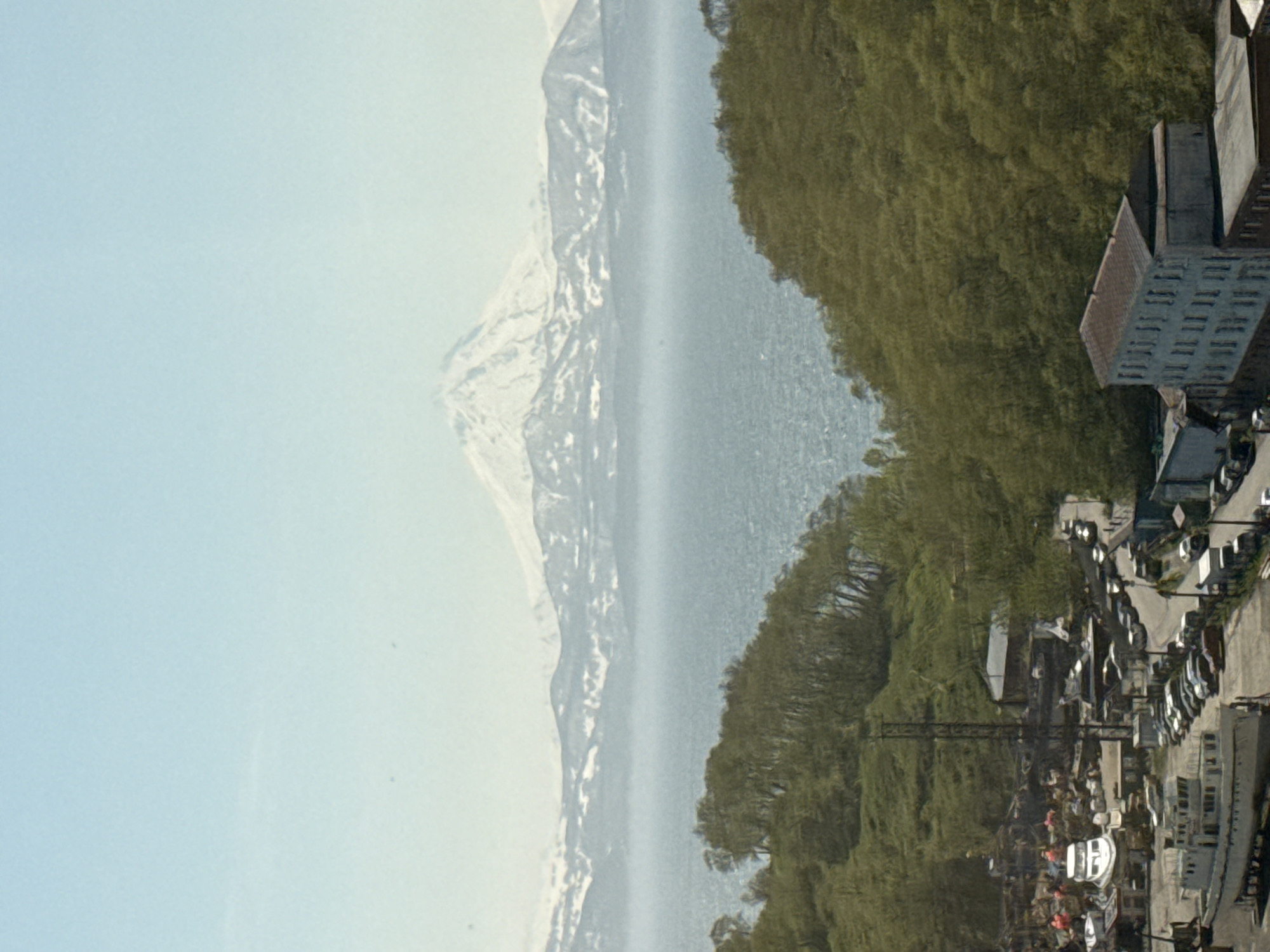 The image depicts a panoramic view of a coastal city with a prominent snow-capped volcano in the background. In the foreground, a densely wooded hillside slopes down towards a harbor area. The harbor is filled with various boats, including several large vessels, and appears to be a hub of activity with several buildings and parked cars. A road winds through the harbor area, lined with more parked vehicles. Further out, the sea stretches towards the horizon, with the majestic, snow-covered volcano dominating the skyline. The sky is clear and pale blue. The scene appears to be a snapshot of daily life in a Russian coastal city, possibly Petropavlovsk-Kamchatsky, given the presence of a volcano and the likely geography.