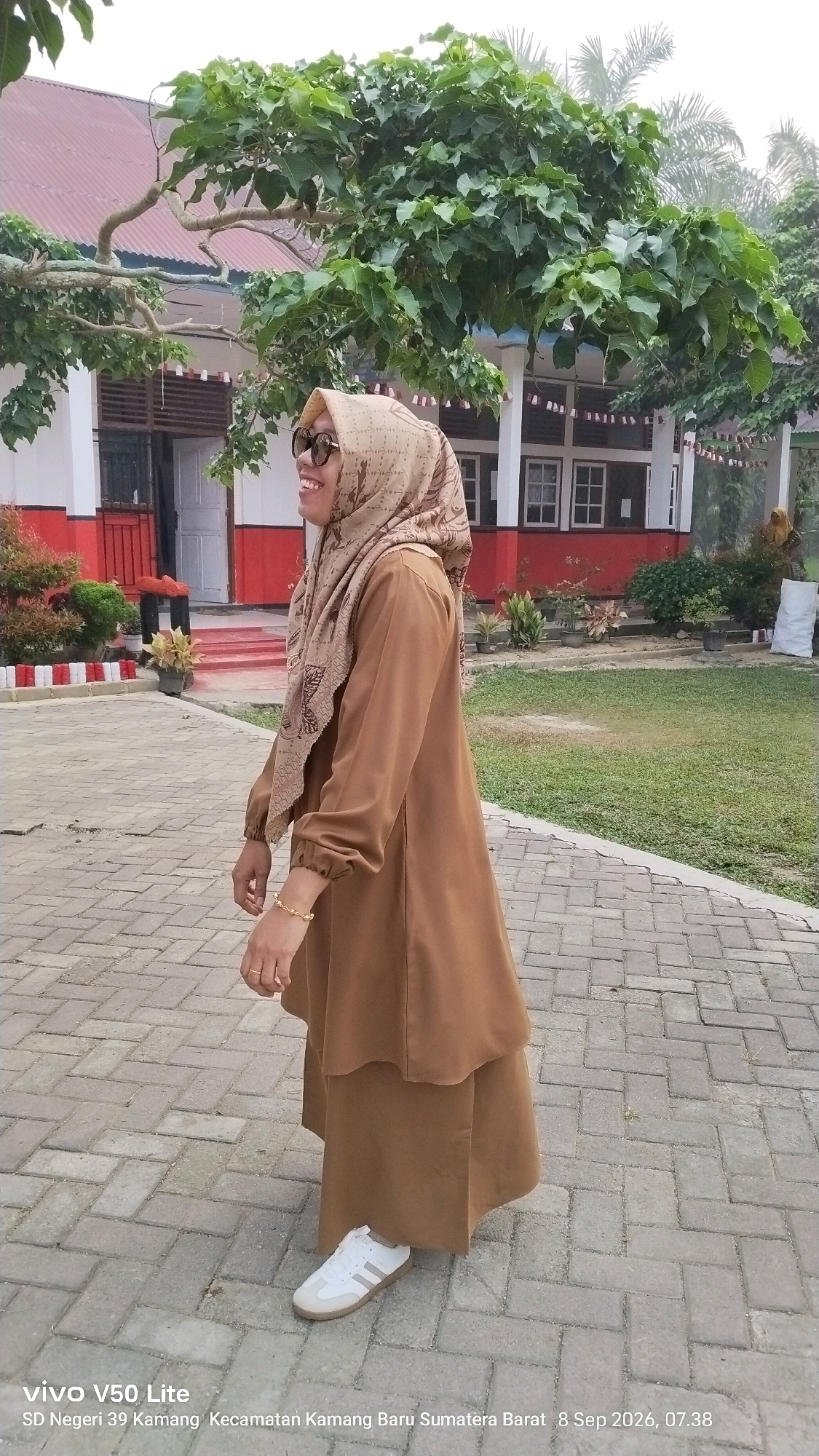 A woman wearing a brown hijab and a matching brown dress stands on a brick pathway outdoors. She is smiling and looking to her left. She wears sunglasses, a gold bracelet, and white sneakers with three grey stripes. Behind her, a building with red and white trim is visible, along with trees and green grass. The sky appears overcast. A watermark in the lower left corner indicates the image was taken with a "vivo V50 Lite" and specifies the location as "SD Negeri 39 Kamang, Kecamatan Kamang Baru, Sumatra Barat" on "8 Sep 2026, 07.38".