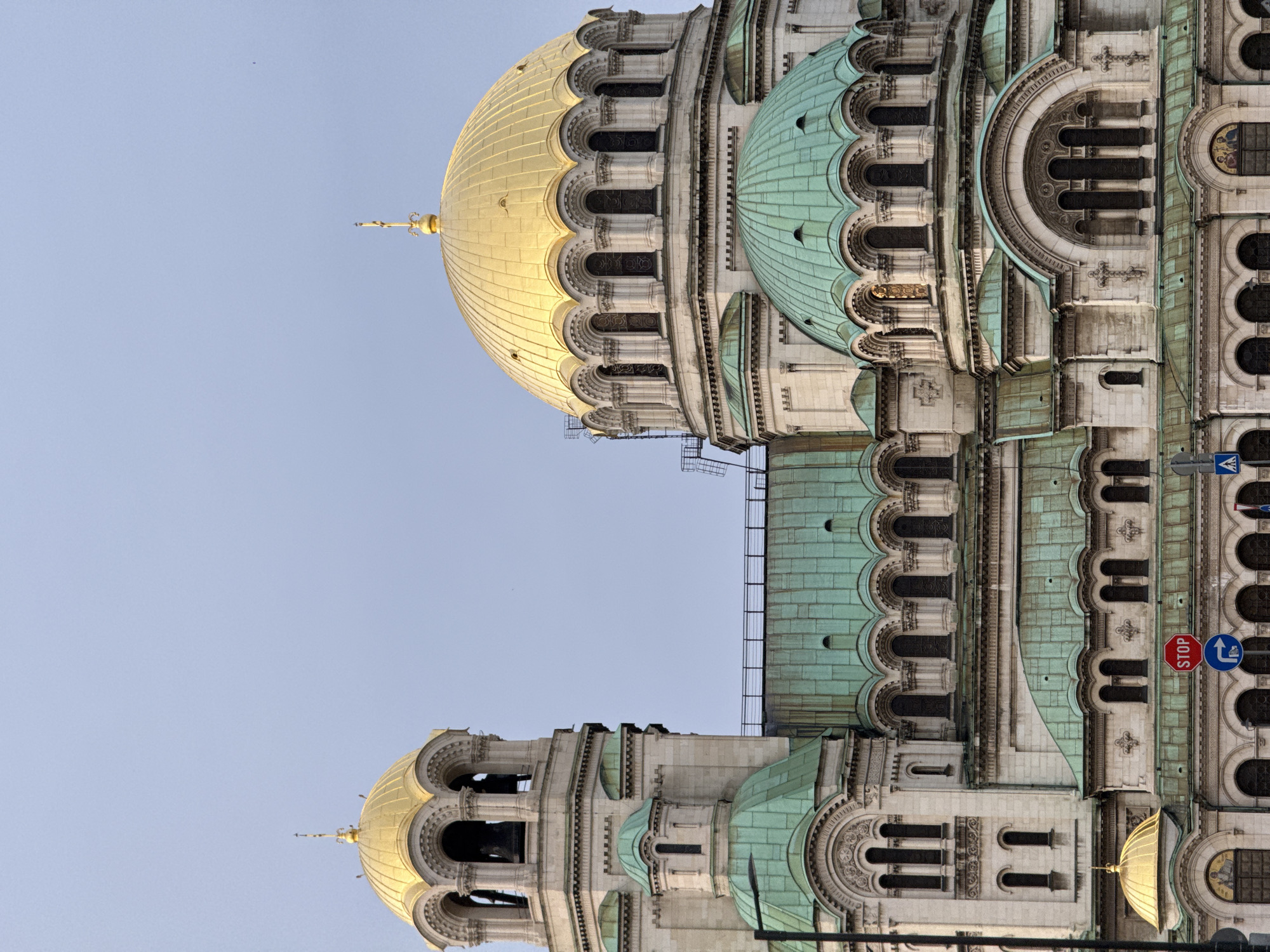 The image depicts the Alexander Nevsky Cathedral in Sofia, Bulgaria. The architectural details of the domes, arches, and decorative elements are visible. A portion of the sky is also present. A stop sign and other traffic signs are visible near the cathedral.