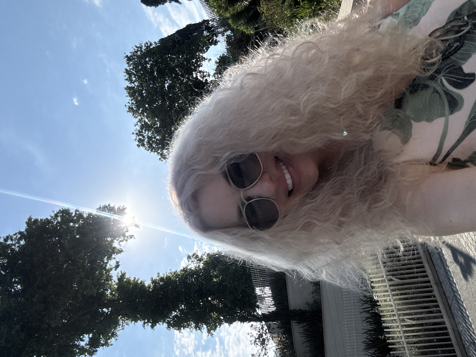 A woman with long, curly blonde hair and sunglasses smiles at the camera, holding it out for a selfie. She is wearing a light-colored dress with a floral pattern in shades of green and pink. The background is a bright, sunny sky with some clouds and lush green trees. In the foreground, a white fence runs along a paved pathway. The image appears to have been taken outdoors in a park or garden setting, potentially in the city of Sochi, Russia, given the Mediterranean-like vegetation and architecture.