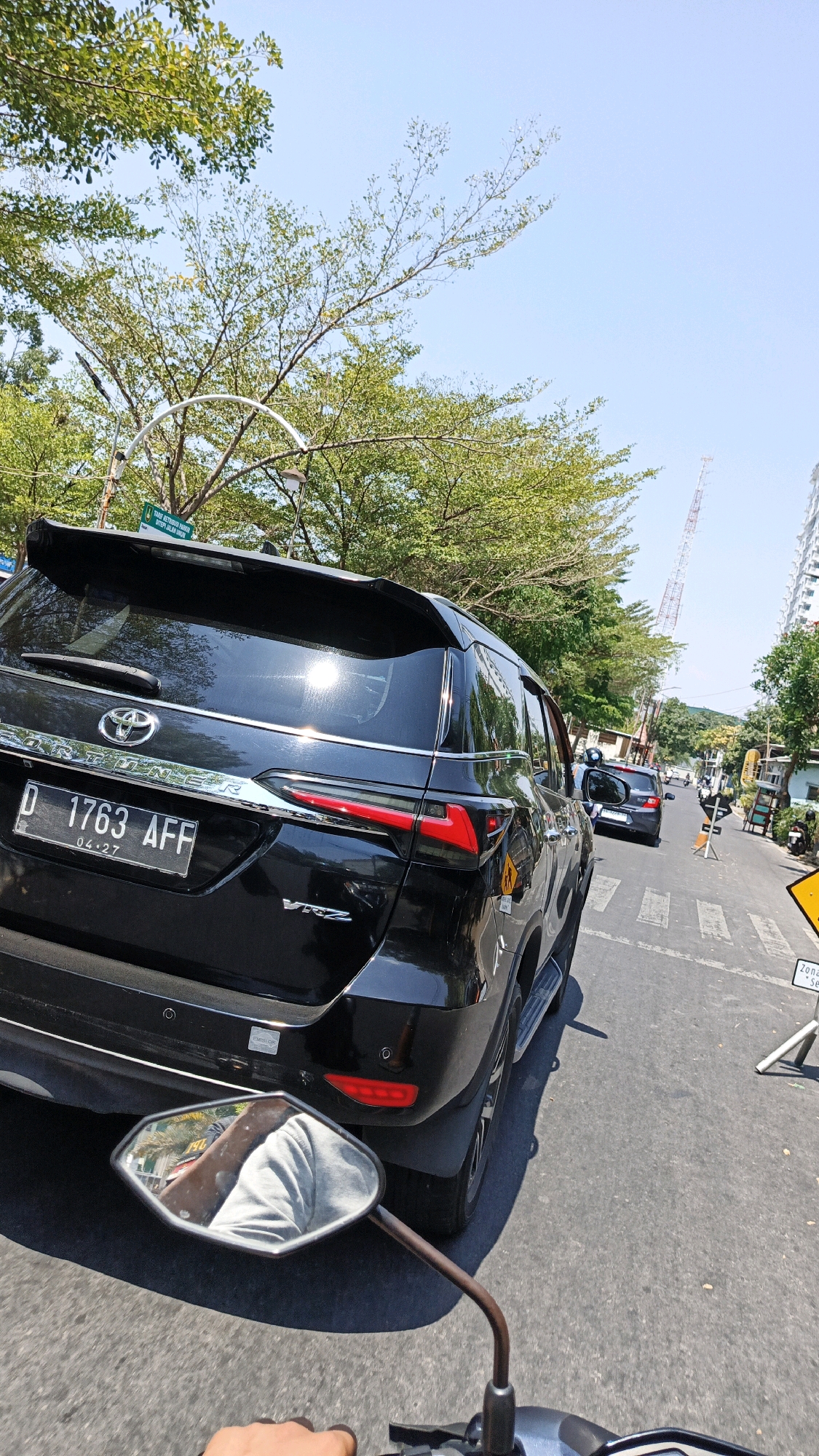 The image, taken from the perspective of a motorcycle rider, shows a black Toyota Fortuner SUV in traffic on a sunny day in Surakarta, Indonesia. The SUV's license plate reads "D 1763 AFF" with an expiration date of "04.27". A reflection of the rider, wearing a grey shirt, is visible in the motorcycle's side mirror. In the background, lush green trees line the street, and a tall transmission tower is visible in the distance. Buildings and other vehicles, including a dark-colored car behind the SUV and several motorcycles, are also present. Road markings and traffic signs, one yellow and triangular with black text, indicate the urban setting. The sky is clear blue.