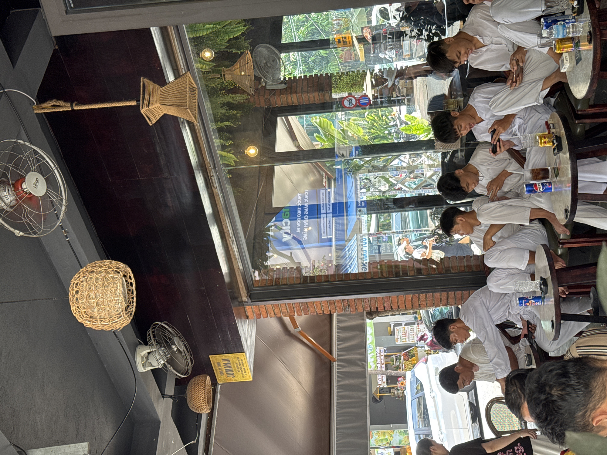 The image depicts the interior of a cafe in Nha Trang, Vietnam. Several young men, dressed in white, are seated at tables, engrossed in their mobile phones. One individual is actively playing a game, with an animated expression. On the tables are drinks, including cans of Pepsi and what appears to be beer, along with ashtrays and cell phones.
The cafe's interior features a dark ceiling with hanging lamps and ceiling fans. The walls are partially covered with brick and wood paneling. Large windows offer a view of the street outside, showing parked vehicles, other shops, and pedestrians. Greenery, likely plants, hangs from the ceiling and is visible through the windows, contributing to a tropical ambiance. A sign in Vietnamese is visible, indicating the establishment's location within the city. The overall scene suggests a casual gathering in a popular urban setting.