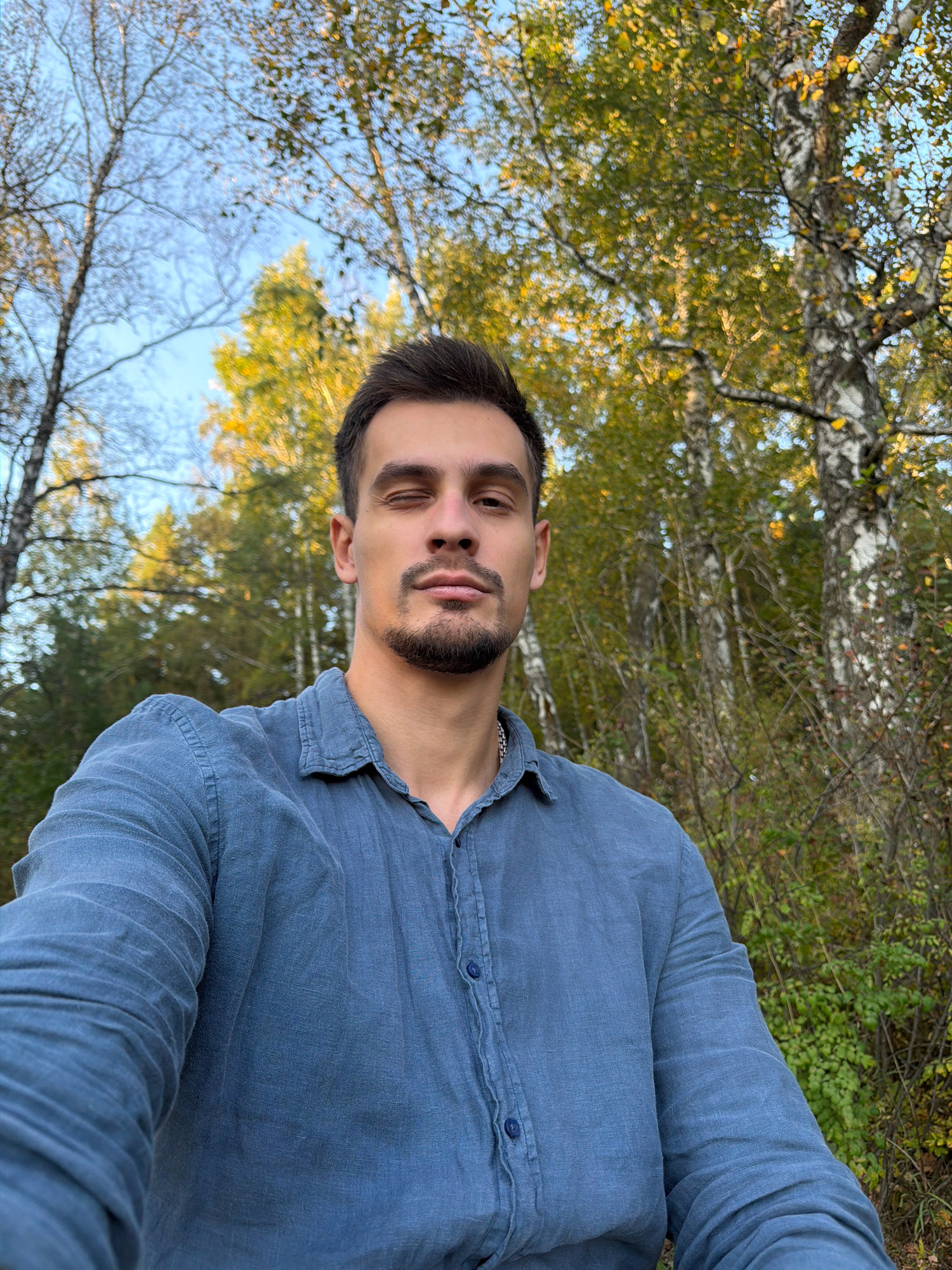 A young man with dark hair and a beard, wearing a blue collared shirt, is taking a selfie outdoors. He is positioned slightly off-center to the left of the frame, with his right eye closed and his head tilted slightly. The background consists of trees with autumn foliage and a bright blue sky. The man's left arm and shoulder are visible in the foreground, extending towards the left edge of the image. The scene appears to be in a natural setting, possibly a park or forest area within the city of Krasnoyarsk, Russia, suggested by the prevalence of birch trees, common in the region. The lighting indicates it is daytime, likely afternoon, given the warm tones of the leaves and the angle of the sun.