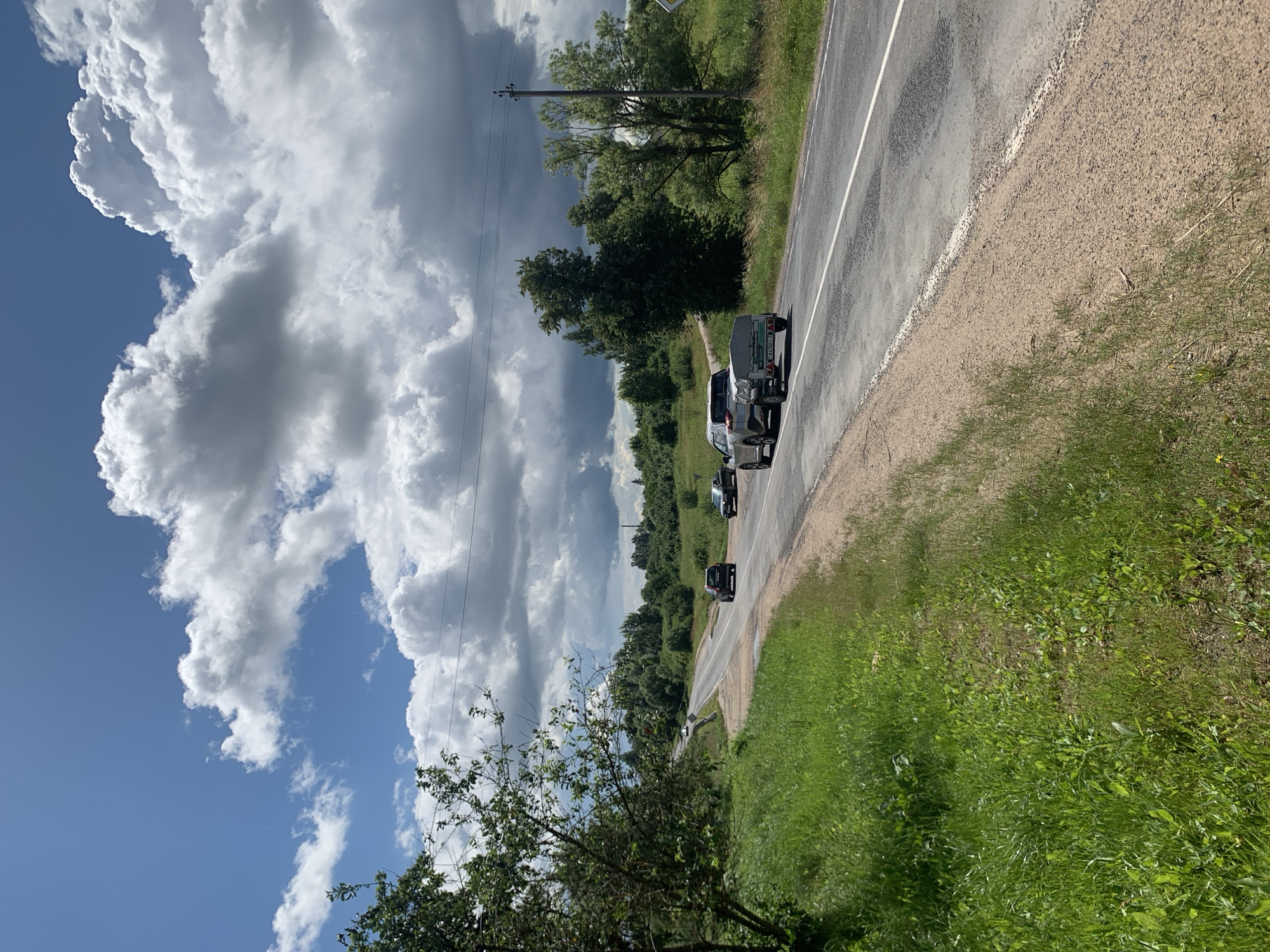 A grey car is towing a small trailer on a two-lane asphalt road that curves to the left, bordered by a gravel shoulder and a grassy verge. The road is flanked by lush green grass and trees on either side. In the distance, the road disappears over a hill, surrounded by more trees and vegetation. The sky is a vibrant blue, dotted with large, fluffy white clouds. The overall scene depicts a rural landscape in Belarus on a bright, cloudy day.
