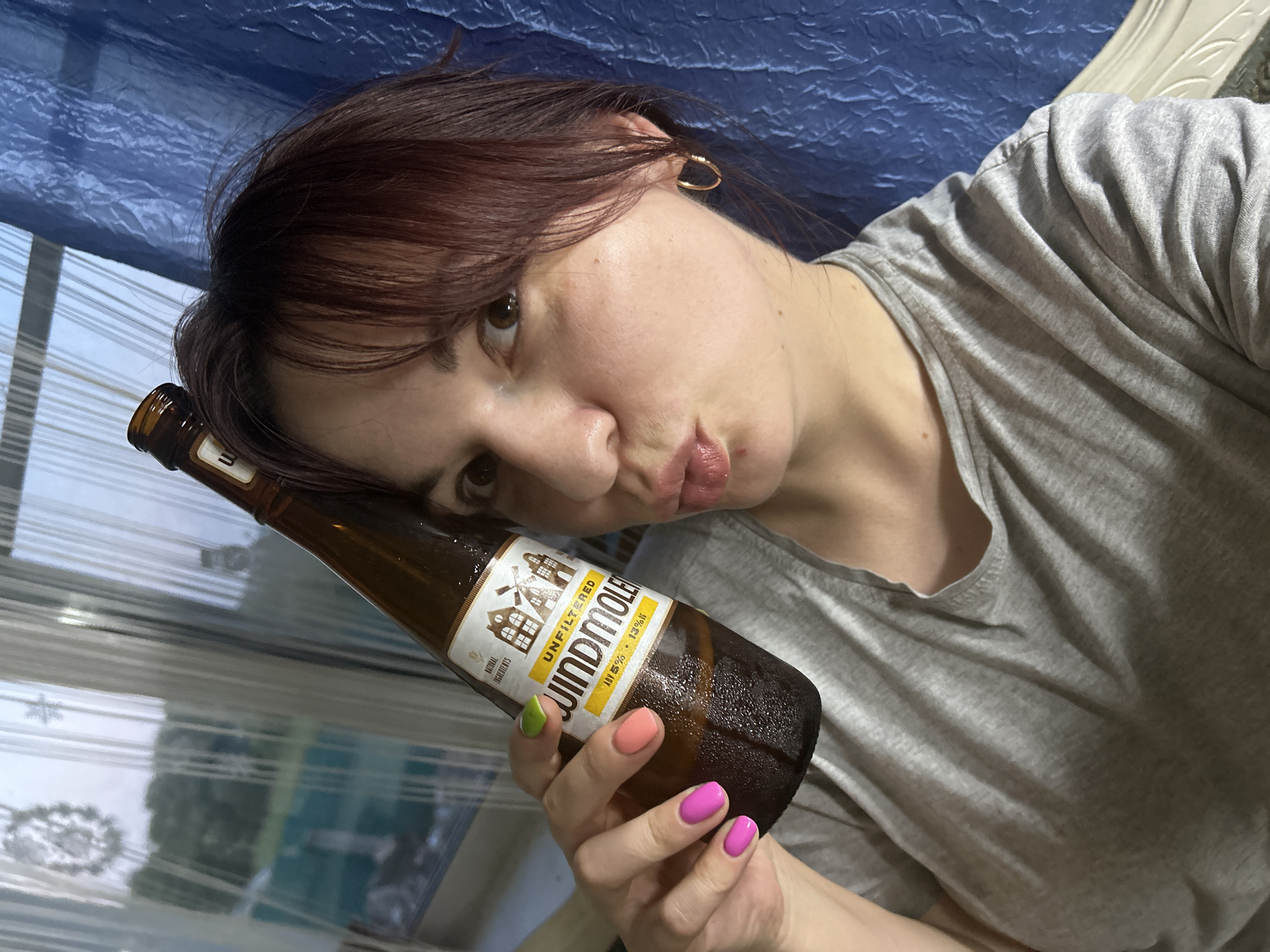 A woman with dark auburn hair and light brown eyes is taking a selfie, holding a bottle of Windmolen Unfiltered beer in her right hand. She is wearing a grey t-shirt and has brightly colored fingernails in pink, orange, and green. Her lips are puckered, and she is looking at the camera. Behind her, a blue textured fabric is visible, along with a window with sheer curtains and a glimpse of greenery outside, suggesting an indoor setting possibly in a residential area. The bottle has a label with an illustration of windmills and text that appears to be in Dutch. There are water droplets on the bottle, indicating it is cold.