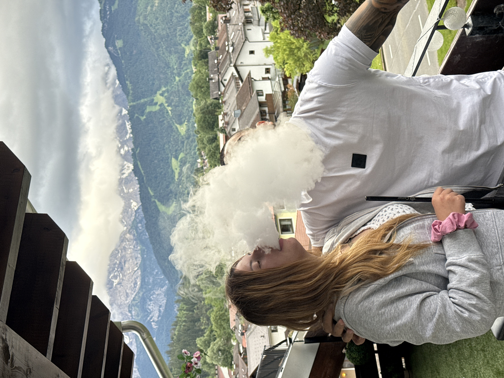 A couple stands on a balcony overlooking the city of Garmisch-Partenkirchen, Germany. The woman on the left, with long brown hair, wears a grey hoodie and a patterned top. She is exhaling a large plume of vapor. The man on the right, wearing a white t-shirt with a black logo on the chest and tattooed arms, has his arm around her. They are positioned in front of a backdrop of lush green mountains partially obscured by clouds. The balcony railing is visible, along with some decorative lights. The background features typical Bavarian-style houses with red-tiled roofs. The image captures a moment of shared activity on a cloudy day with a scenic mountain view.