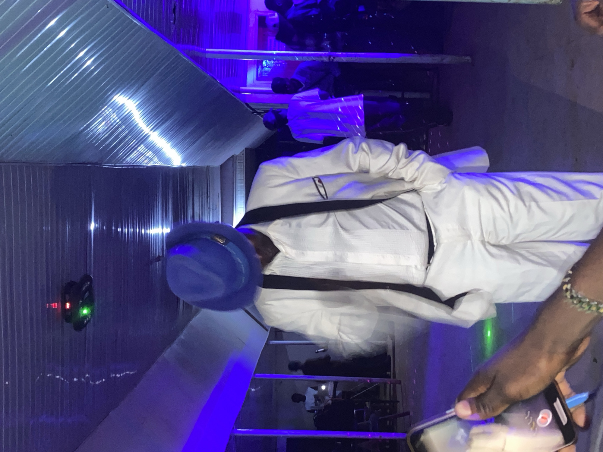 In an indoor setting illuminated by blue and white lights, a person dressed in a white suit and a blue fedora stands in the foreground, facing away from the camera with their head bowed. The person's right hand is raised, and their left hand is partially visible in the lower left corner, holding a smartphone that is recording. The environment appears to be a venue with a corrugated metal ceiling and tiled walls, possibly a community hall or a large room. Other individuals are visible in the background, blurred by the low light and movement. A green laser light is projected on the floor in the middle ground. The overall impression is a lively event, possibly a party or a performance.