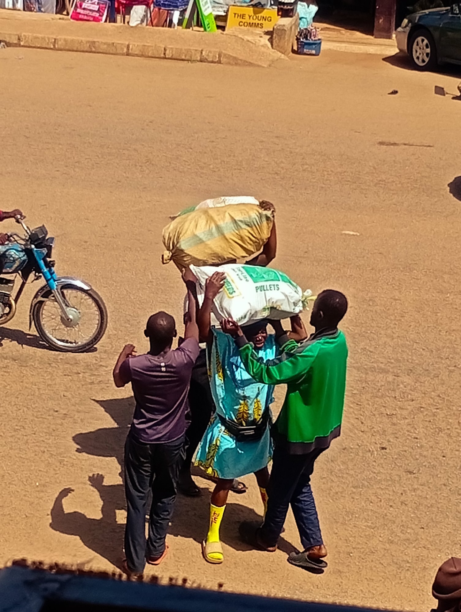 In Bukuru, Nigeria, four men are carrying a large, yellow sack and a white sack labeled "PULLETS" down a sandy, sun-drenched street. The man on the far left, wearing a purple shirt and dark pants, appears to be guiding the load. To his right, a man in a turquoise shirt is supporting the white sack. A man in a green jacket, on the right, also helps carry the sacks. Another man, mostly obscured by the sacks, is also involved in carrying them. In the background, a blue motorcycle is parked, and market stalls are visible. A dark-colored car is parked further down the street. The ground is a mix of sand and dirt, with shadows indicating a bright, sunny day.