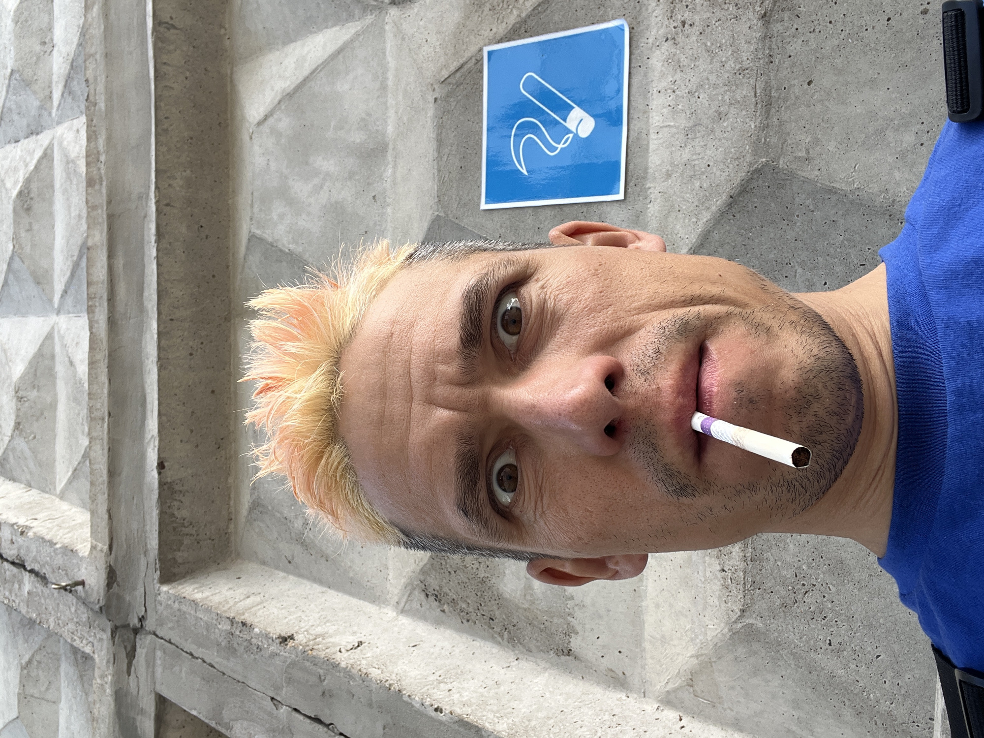 A fair-skinned male with blonde-tipped hair and a stubble beard is holding a cigarette in his mouth. He is wearing a blue shirt and standing in front of a concrete wall. A blue sign with a white cigarette graphic is affixed to the wall above his head. The wall appears to be part of an architectural structure composed of repeating geometric patterns. The scene is in an outdoor location and appears to be in the city of Chaikovsky, Russia.