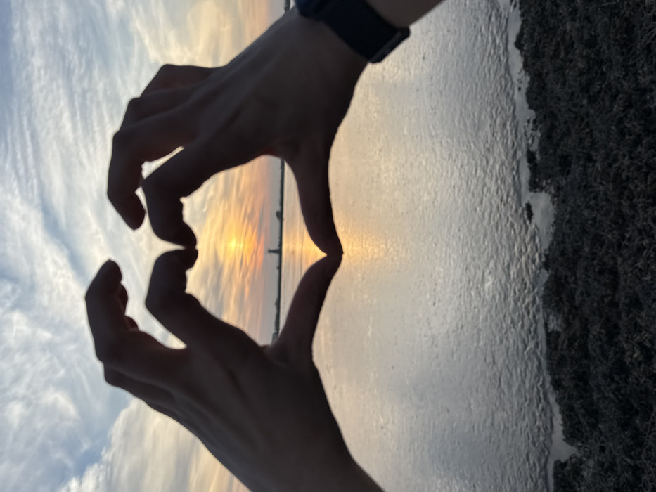 This image depicts a sunset over a body of water, likely Breydon Water or the River Yare, located in the vicinity of Great Yarmouth, United Kingdom, with a view looking westward. In the immediate foreground, two human hands, belonging to a single individual, are silhouetted against the bright horizon, forming a heart gesture. A dark-colored smartwatch is visible on the wrist of the right hand. The setting sun, a bright orange-yellow disc, is partially framed within the heart shape formed by the hands. The midground features a wide expanse of calm water, reflecting the warm light of the sunset. The bottom portion of the image displays a dark, textured waterline, consistent with marshland vegetation or accumulated debris along a bank. On the distant horizon, silhouetted against the radiant sky and sun, a tall, slender tower structure is visible, consistent with the Berney Arms Windmill, a known landmark west of Great Yarmouth. The sky above is streaked with wispy clouds, illuminated in shades of orange, yellow, and blue.