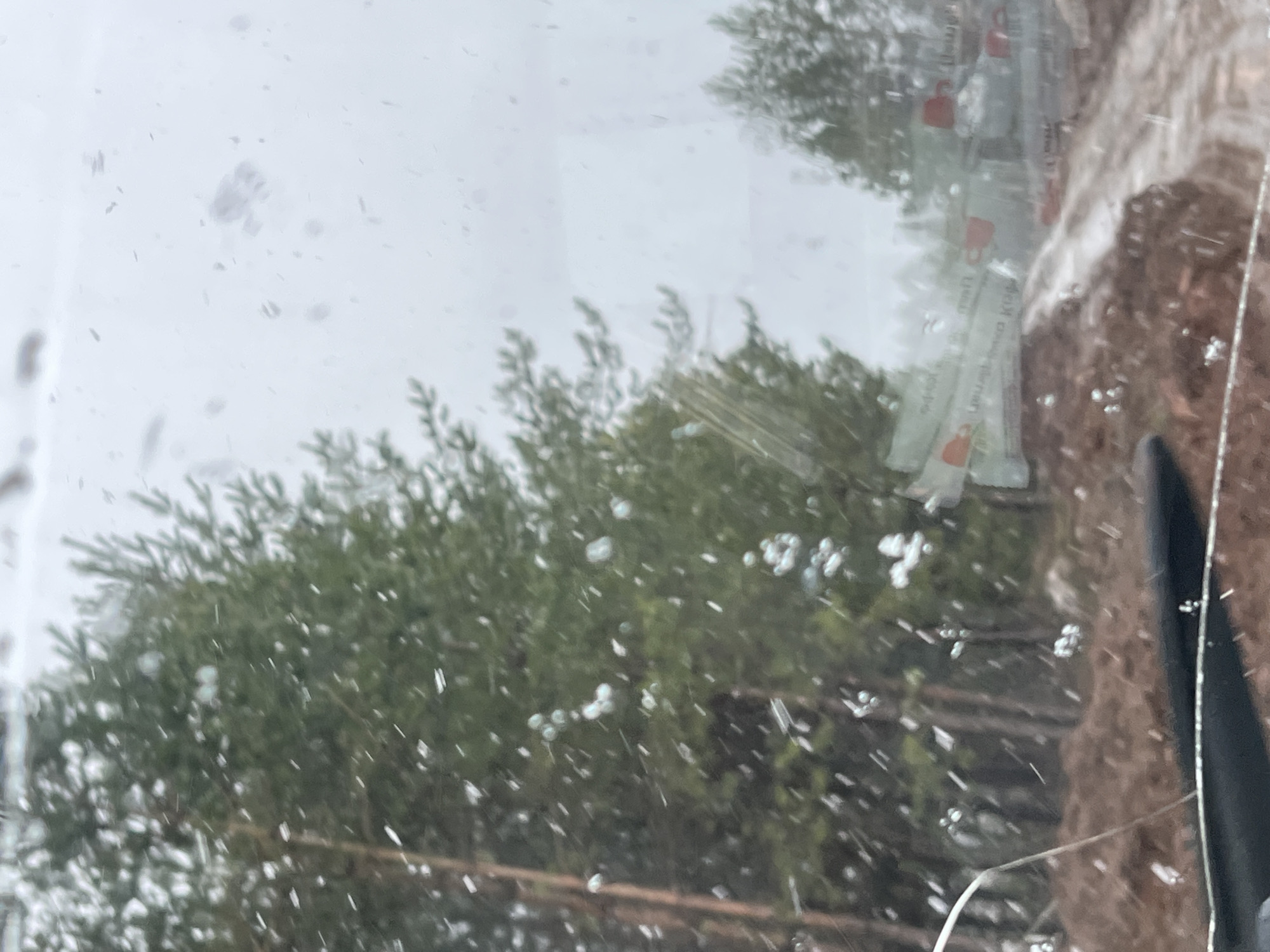 The image, taken through a window, shows snow falling. Outside the window are green trees and part of a building with stacked coffee creamer packets affixed to it. Cracks are visible in the lower right corner of the window. Given the mention of Chaikovsky, Russia, this scene is presumed to be located there.