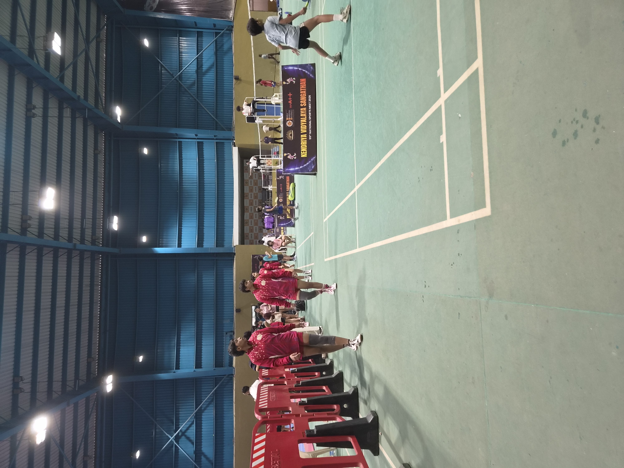 The image depicts the interior of a gymnasium, likely hosting a sporting event, possibly badminton or volleyball, as indicated by the court markings. In the foreground, two young individuals wearing matching maroon athletic outfits and knee pads are walking across a light green court. To the left, a red barrier with white stripes separates the court from a walkway. In the background, several other individuals, some in athletic wear, are visible, including a person seated on a high umpire's chair. A large banner with text "KENDRIYA VIDYALAYA SANGATHAN 55th NATIONAL SPORTS MEET 2026" is displayed on a dark background. The gymnasium has a high, corrugated metal roof supported by blue beams, and illuminated by overhead lighting fixtures. The overall environment suggests an organized sports competition within an indoor facility. The scene is located in Phagwara, India.