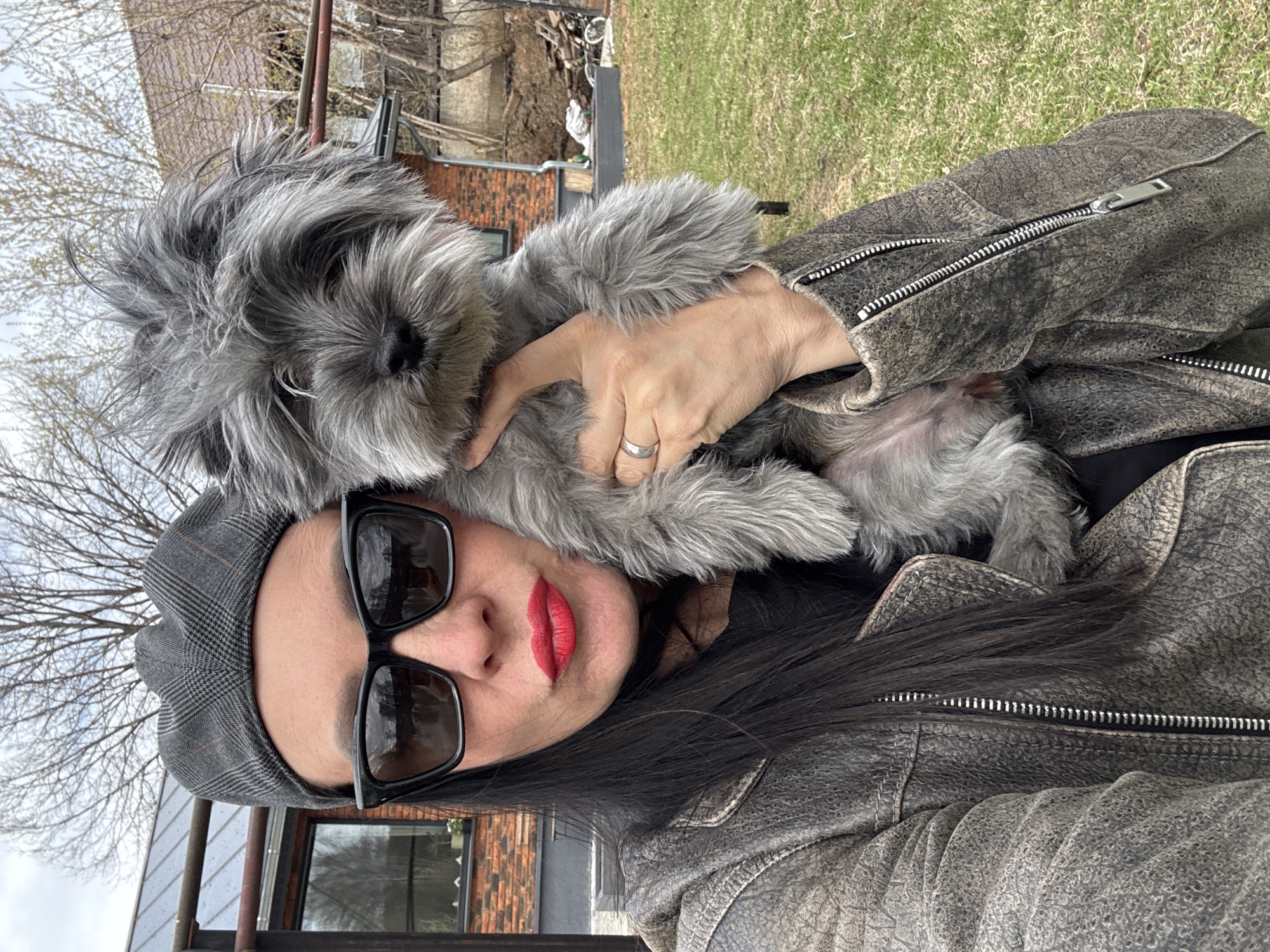 A woman is holding a grey, fluffy small dog. She is wearing a grey plaid hat and large black sunglasses and bright red lipstick. She is wearing a textured grey jacket. The background includes a brick building, a green lawn, and bare trees. The image was taken in Chaikovsky, Russia.