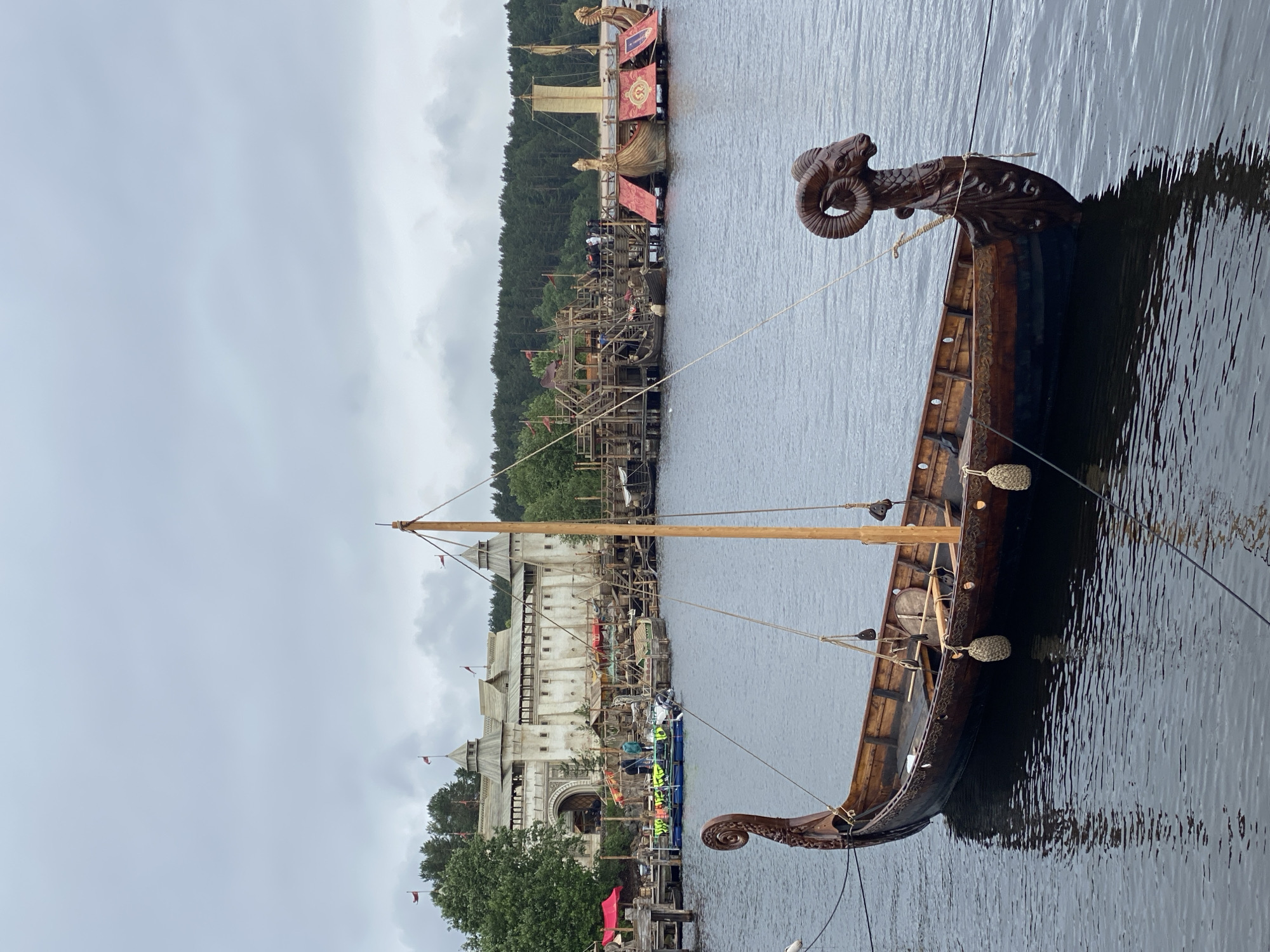 In the city of Моторное, Russia, on a cloudy day, a Viking longship with a carved ram's head prow is moored in a body of water. The ship is dark wood with intricate carvings along its hull and is equipped with a single mast and rigging. In the background, a large, fortified wooden structure resembling a castle or settlement sits on the shore, with several smaller boats and platforms visible around it. The sky is overcast, and the water is calm, reflecting the surrounding structures and the ship. The scene suggests a historical reenactment or a themed attraction.