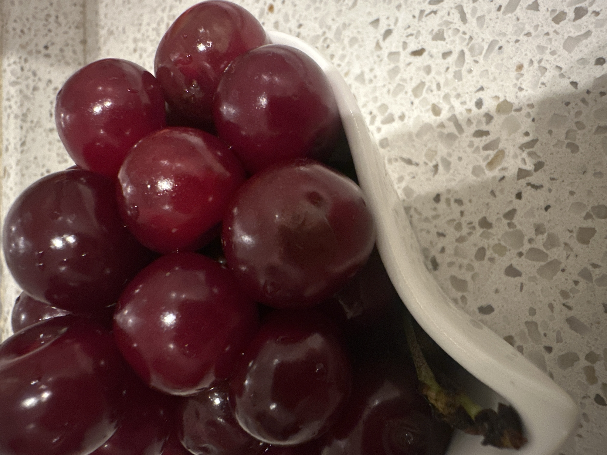 A close-up, overhead shot shows a pile of dark red cherries in a white bowl. The cherries are plump and glossy, with some appearing to have water droplets on their surface. The bowl is positioned on a speckled countertop that is light gray with white and brown flecks. The background is slightly blurred, but appears to be a kitchen environment. The cherries are the primary subject of the image, and they fill most of the frame. The lighting in the image is bright and diffused, creating highlights on the cherries and making their color appear rich and vibrant.