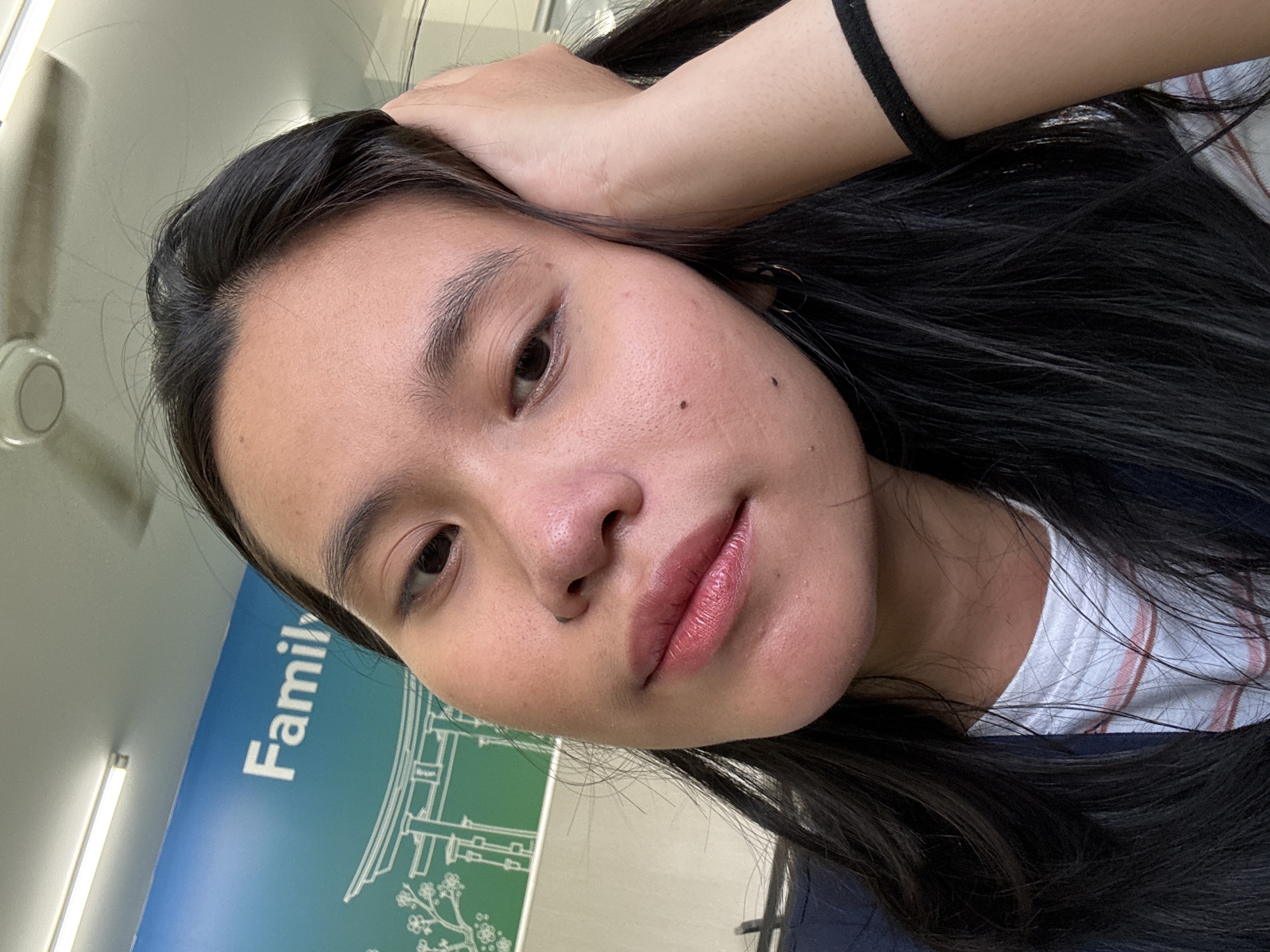 A young woman with long, dark hair is captured in a selfie, her head tilted slightly as her right hand rests on her head. She is wearing a dark jacket over a white t-shirt with thin pink and white stripes. A black band is visible on her right wrist. Her lips are colored with a shimmery pink lipstick. In the background, a blue wall with white lettering that reads "Family" and an illustration of a traditional Japanese gate and cherry blossoms is visible. The ceiling above shows fluorescent lights and a fan. The scene appears to be indoors, possibly in a public space or office in East Jakarta, Indonesia.