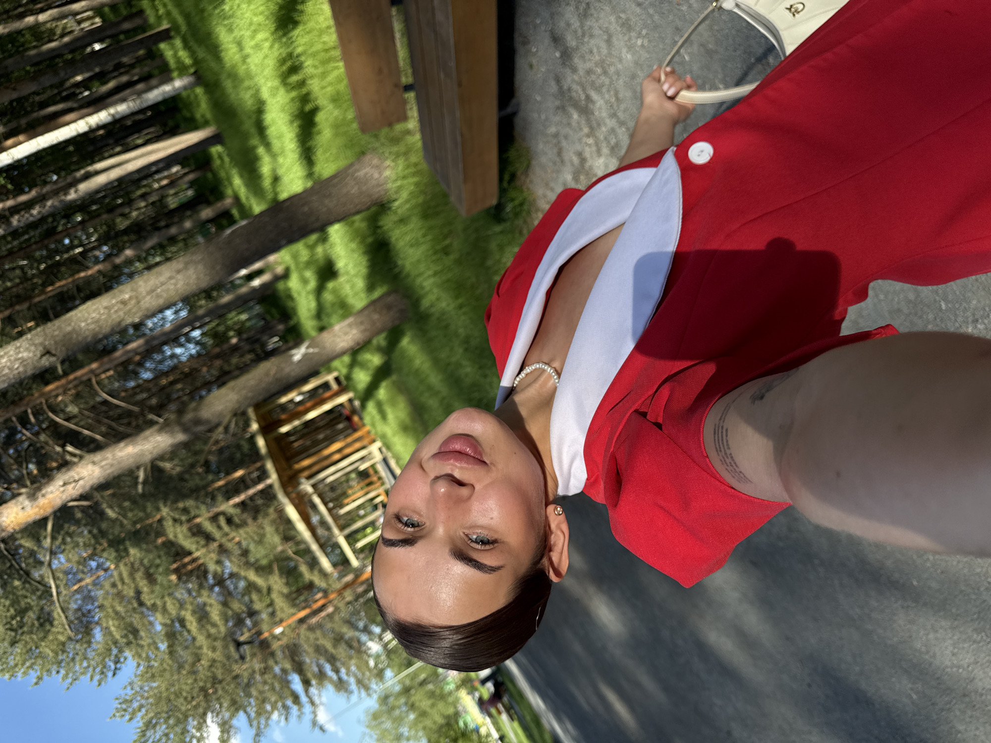 A woman is standing outdoors near a park in what appears to be Perm, Russia. She wears a red blazer with a white collar, a pearl bracelet, and earrings. She holds a white purse. In the background, there are trees, green grass, a wooden bench, and a structure resembling a playground or treehouse. The area is paved and appears to be a walkway or street within the park. The sky is blue with some clouds visible through the trees. The photo is taken at an angle, likely as a selfie.
