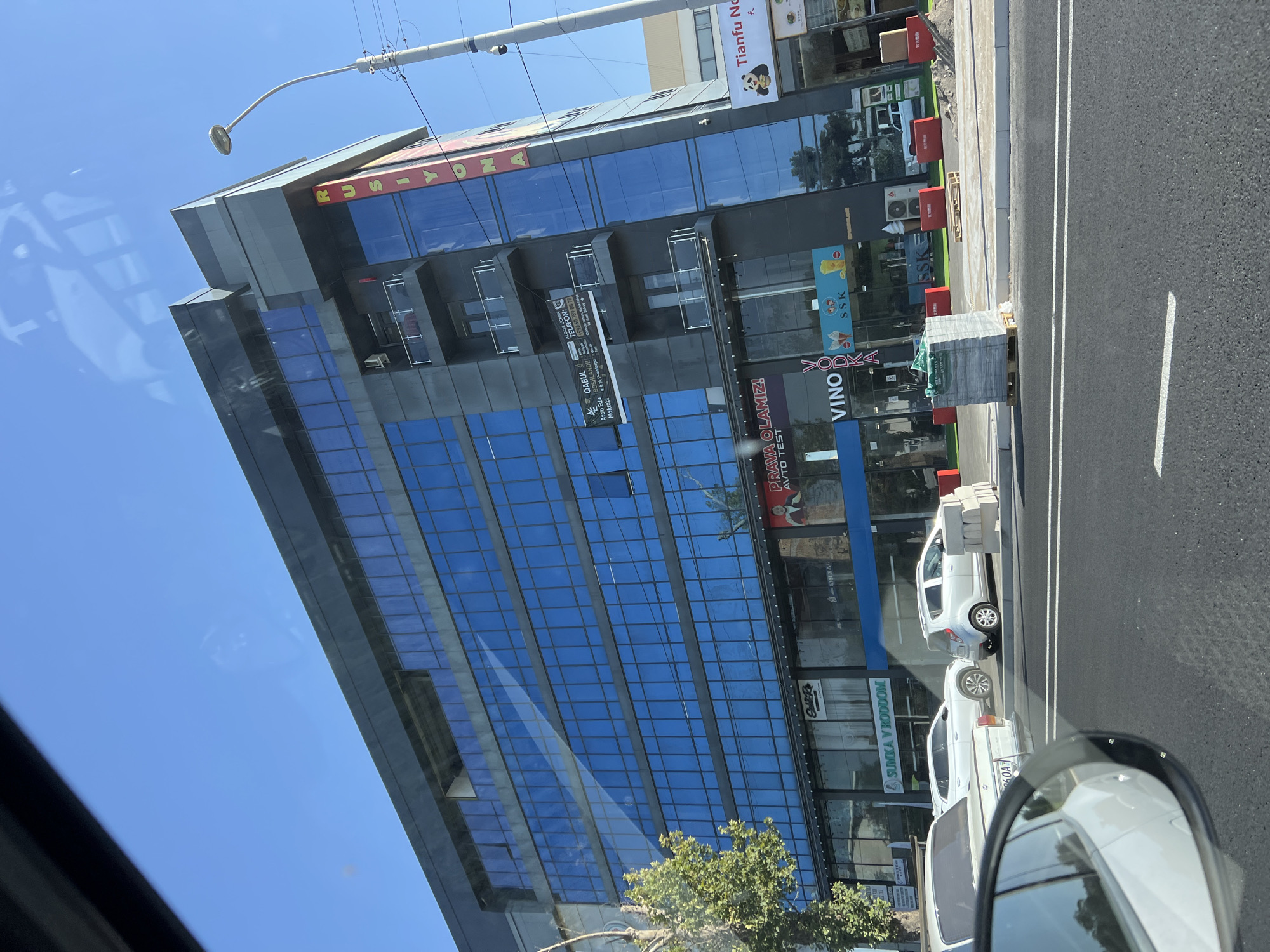 A contemporary, multi-story commercial building with a dark facade and extensive blue-tinted glass windows dominates the frame under a clear blue sky. The building features multiple balconies with glass railings. Prominent signage in vertical red and yellow letters on the side of the building reads "RUSİYONA". At street level, various businesses are visible, including "VINO", "VODKA", and "SSK". Several white cars are parked or driving in front of the building, with one partially visible in the foreground, reflected in a car's side mirror. Construction materials, including stacked bricks and wooden pallets, are present on the sidewalk. A street lamp with a modern design is situated to the right of the building. The overall environment suggests an urban setting.
