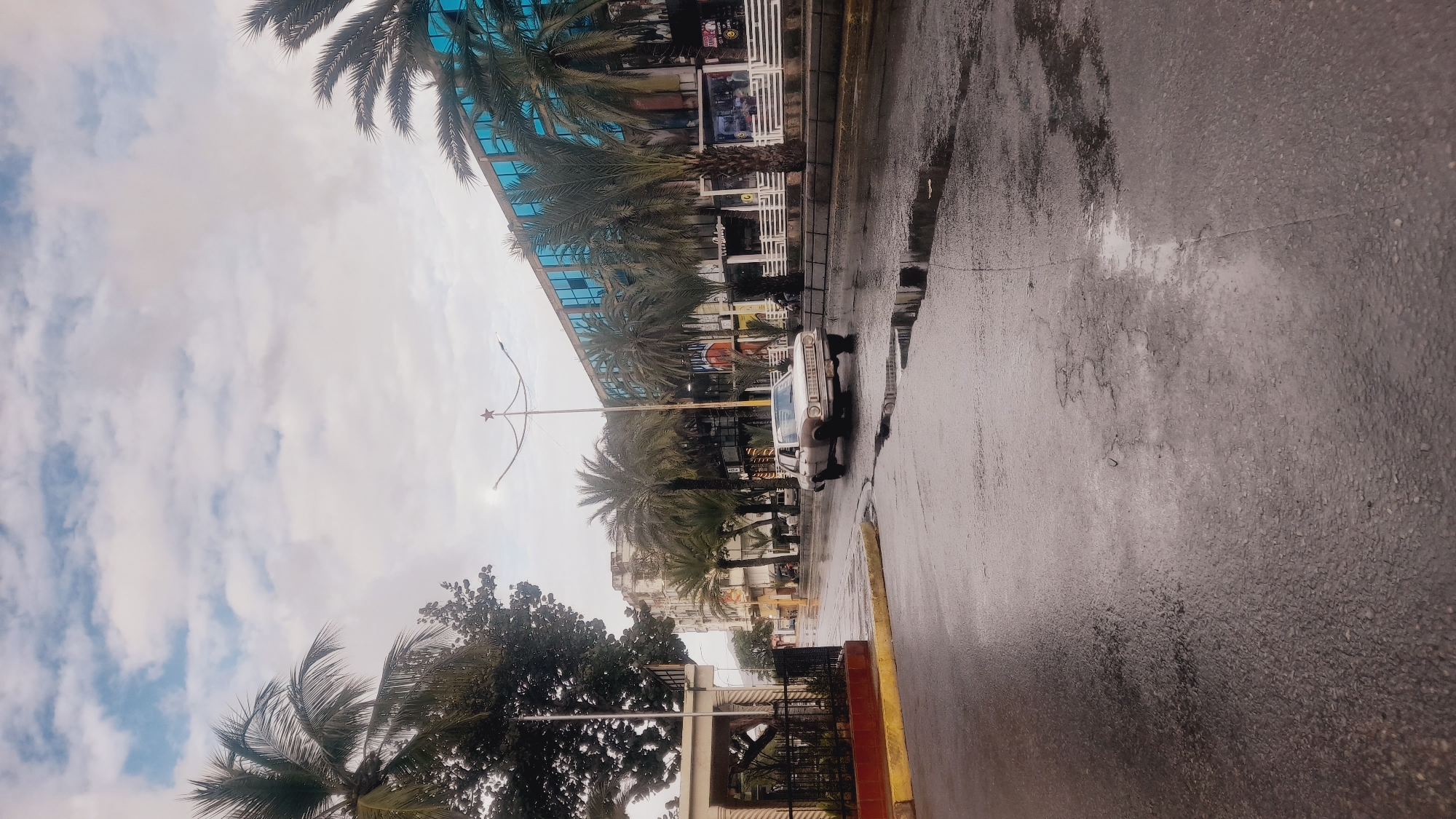 This media file depicts a street scene in Juan Griego, Venezuela, likely after a rainfall. The image is oriented sideways. The road is wet and reflective, with puddles indicating recent precipitation. A white sedan is parked on the right side of the road, facing away from the viewer.
The background features a modern building with blue-tinted glass windows and a facade adorned with white horizontal slats. Palm trees line the street in front of the building, their fronds contrasting with the architecture. Further down the street, other buildings of varying heights and architectural styles are visible.
A street lamp with a star-shaped decoration stands on the left side of the road, extending over the street. The sky is overcast with scattered clouds, suggesting a cloudy or potentially intermittently rainy day. The overall atmosphere appears somewhat subdued due to the weather conditions.
No people are clearly visible in the frame. The image captures a moment of quietude on a wet urban street. No discernible text is visible in the image.