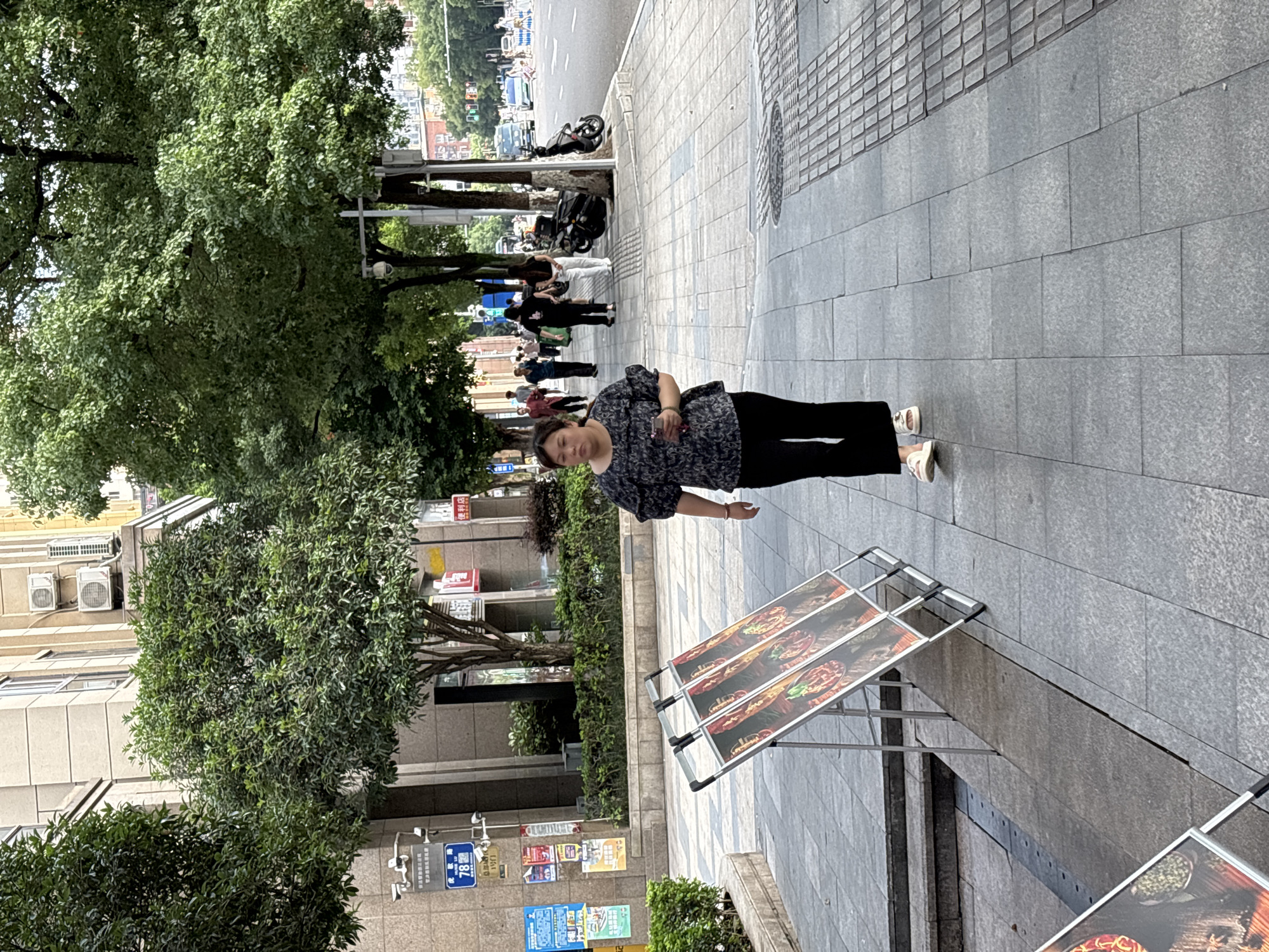 A woman is walking on a sidewalk in the city of Yuzhong District, China. She is wearing a dark patterned shirt and black pants. There are several trees lining the sidewalk, and buildings can be seen in the background. There are also several signs on display, some of which appear to be advertisements. A road with vehicles is visible to the right of the sidewalk.