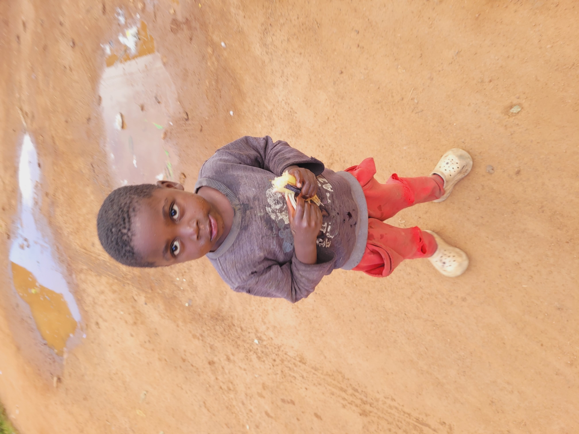 A young boy with dark skin and short, dark hair stands on a dirt road in Bukuru, Nigeria. He is wearing a long-sleeved gray shirt with a graphic print and red pants with large pockets. On his feet, he wears white clogs. He is holding a piece of fruit, possibly a banana, in both hands, which he is in the process of eating. The dirt road is reddish-brown and appears to be slightly muddy in places, with several puddles reflecting the sky. The background is blurred, but suggests an outdoor, undeveloped environment.