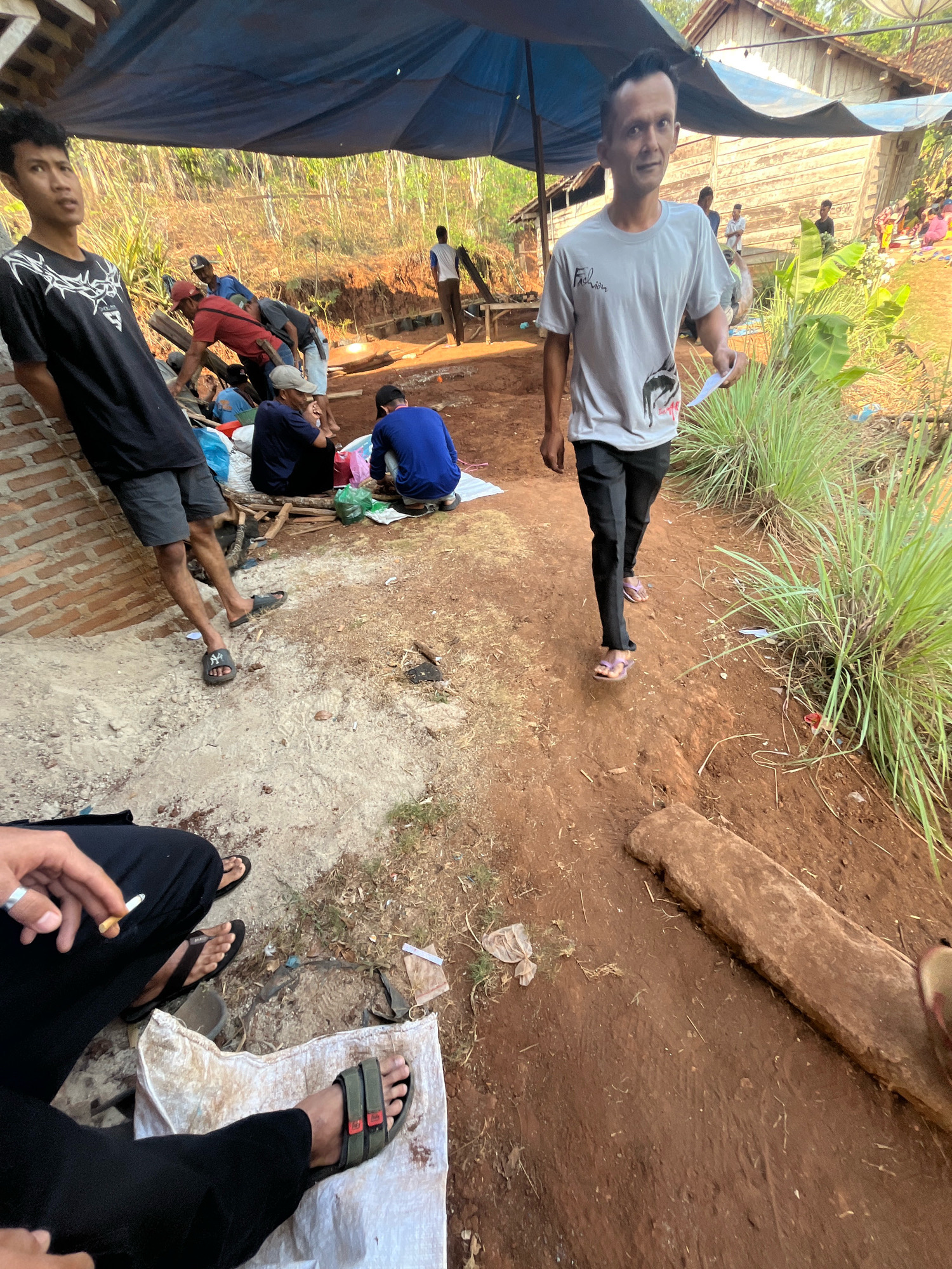 In the city of Sidomulyo, Indonesia, a group of individuals are gathered outdoors, possibly at a construction or communal work site. The scene appears to be during daylight hours with natural lighting.
In the foreground, a person is partially visible, seated with their legs extended. They are wearing black pants and sandals and are holding a lit cigarette, with a silver ring on their left hand. Another person is standing to the left, leaning against a brick wall. This individual is wearing a black t-shirt with a white graphic design and dark shorts, also wearing sandals.
Moving further into the scene, a man in a light grey t-shirt with "Fashion" printed on it, and black pants and sandals, walks towards the viewer, holding a piece of paper. Behind him, a blue tarp is stretched over a wooden frame, providing shade for a group of people. Some individuals are seated or crouched under the tarp. One person wears a red shirt and is actively engaged in an activity. Others are in blue shirts, and some are partially obscured. Several bags, including plastic bags and what appears to be a woven sack, are scattered on the ground near the seated individuals.
The ground surface is a mix of dry, sandy soil and reddish-brown dirt. In the background, a wooden house structure with a visible roof and walls is situated on a slight incline. The surrounding vegetation consists of tall, green grass and dried foliage. The overall environment suggests a rural or semi-rural setting.