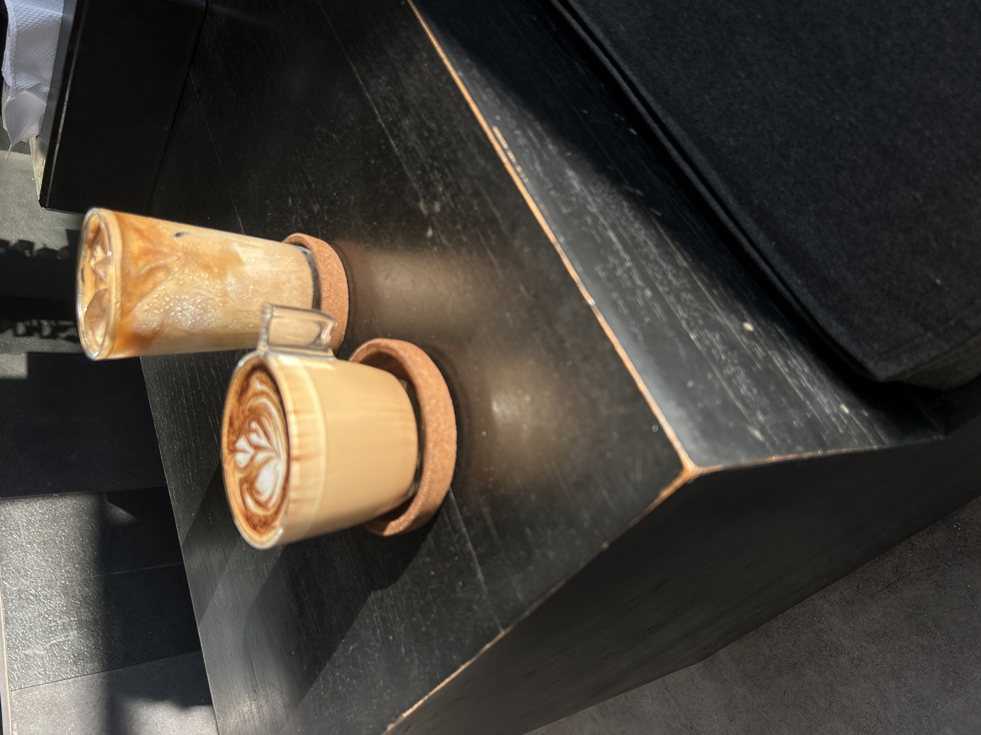 Two coffee drinks are present on a dark, wooden table. The drink in the foreground is a latte in a clear glass mug, topped with latte art, sitting on a cork coaster. The drink in the background is an iced coffee in a tall, clear glass, also on a cork coaster. To the right of the iced coffee is a black tissue box. The table appears to be in an indoor setting, with a dark, textured wall visible in the background, illuminated by sunlight casting shadows. The location is identified as North Kuta, Indonesia.