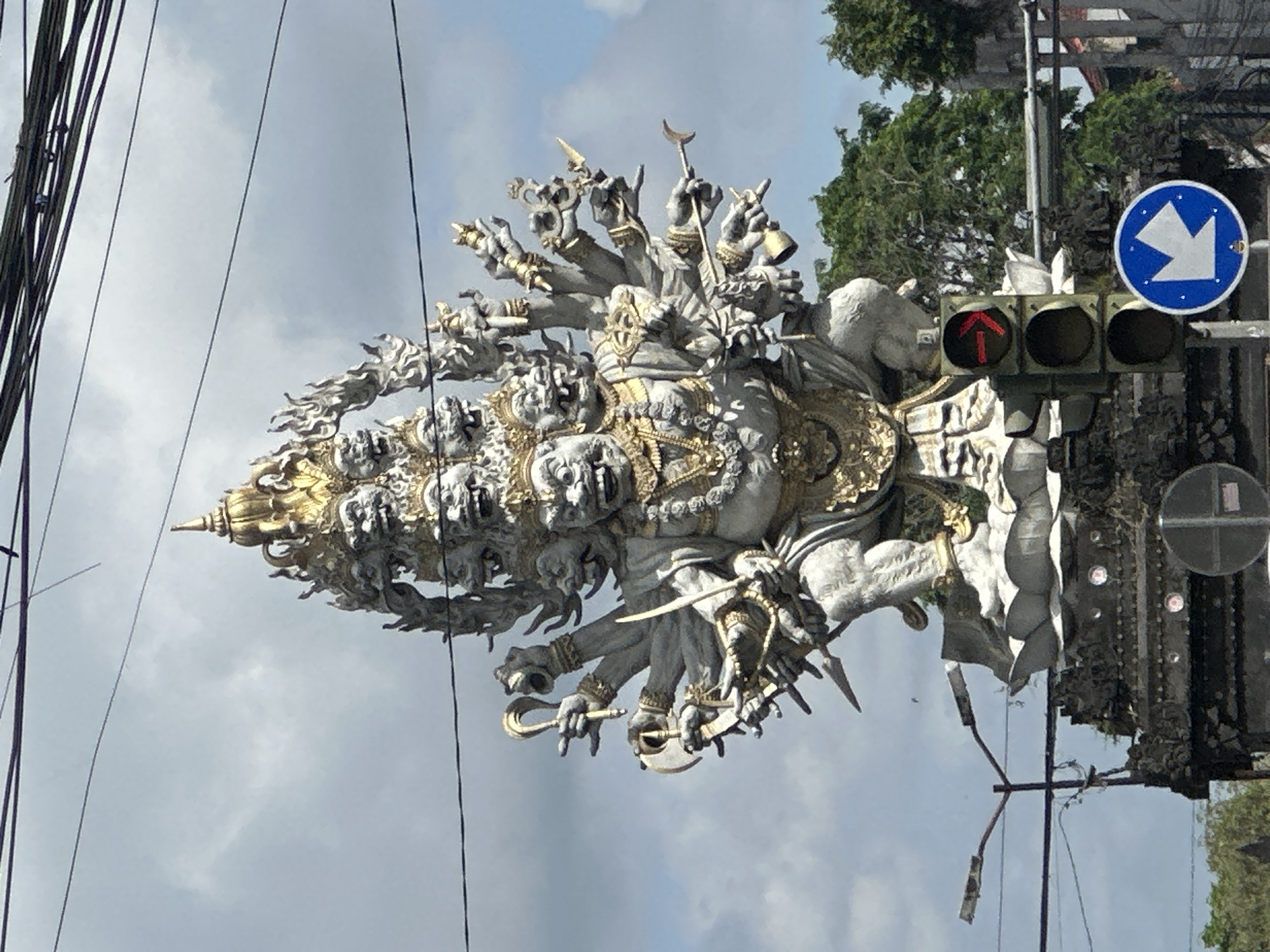 The image shows the "Tugu 9 Naga" monument in Kediri, Indonesia, situated at a busy intersection. The monument is a towering statue of a multi-headed, multi-armed figure, predominantly white with gold accents. The statue stands near a traffic light, which is showing a red arrow indicating that people cannot go straight ahead. A blue and white arrow road sign points to the bottom right of the image. Power lines and lush greenery are visible in the background. The sky is overcast. The photo appears to be taken from a low angle, looking up at the statue.