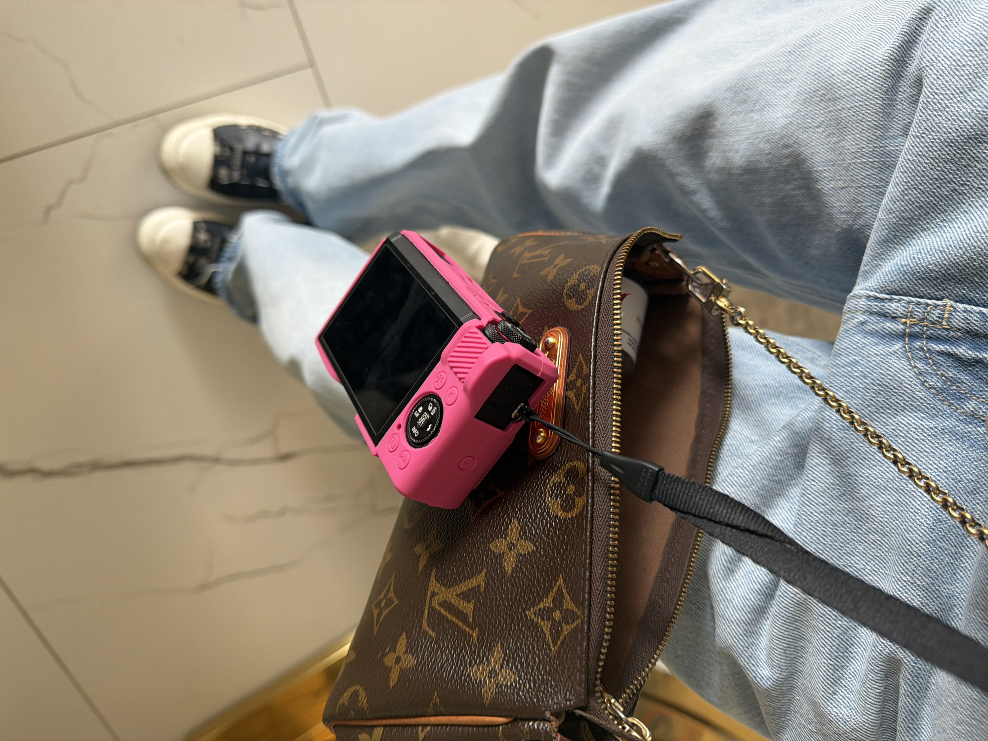 The image depicts a person sitting with their legs crossed, showcasing a fashionable ensemble. They are wearing light blue denim jeans and black and white sneakers. A brown Louis Vuitton handbag rests on their lap, with a small pink camera placed on top of it. A black strap is connected to the camera and drapes over the bag. The background consists of a light-colored tiled floor with a subtle marbled pattern. There is no specific information in the image that points to the location being in Krasnoyarsk, Russia.